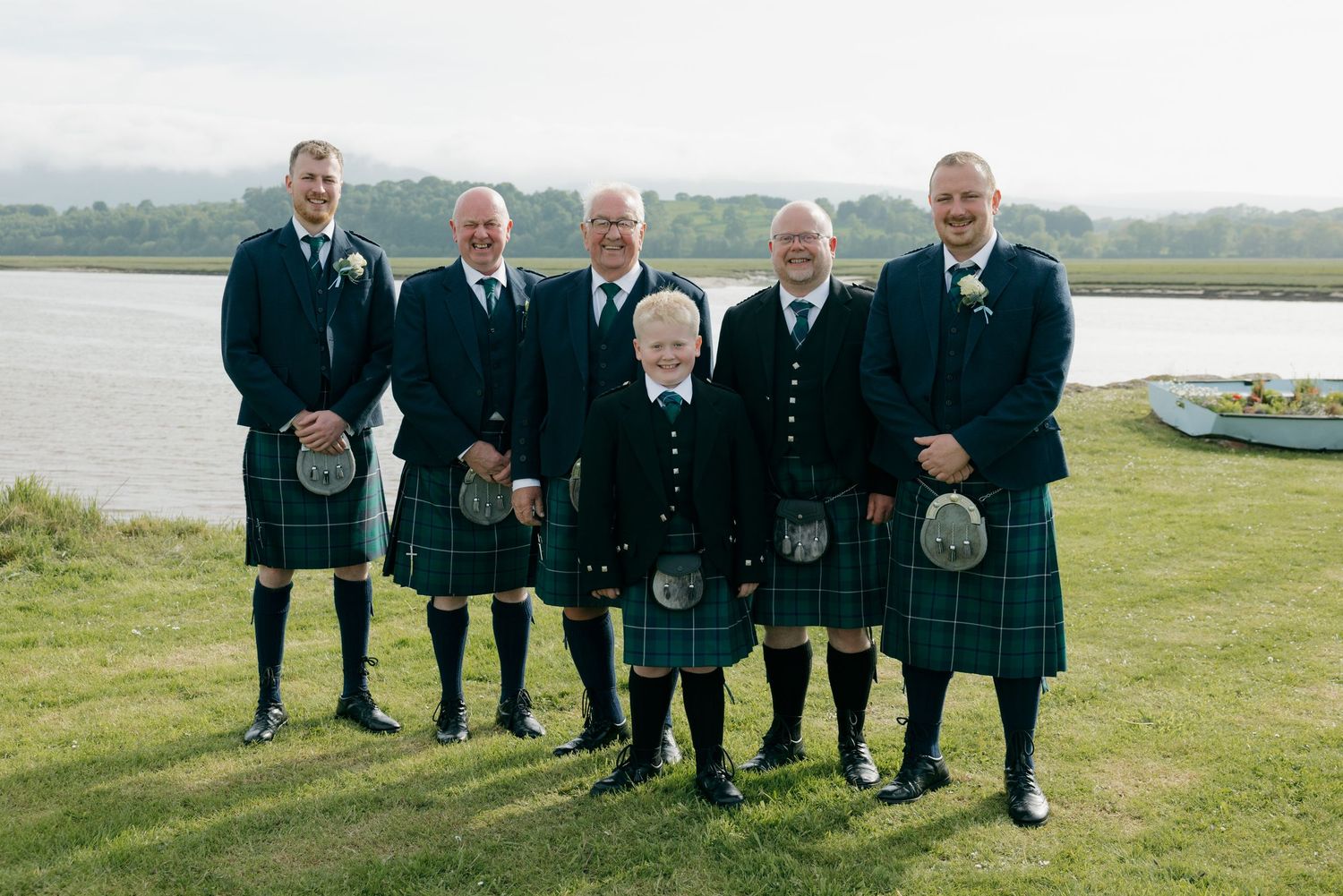 Six men in matching blue tartan kilts pose outdoors near a loch at a Scottish wedding celebration.