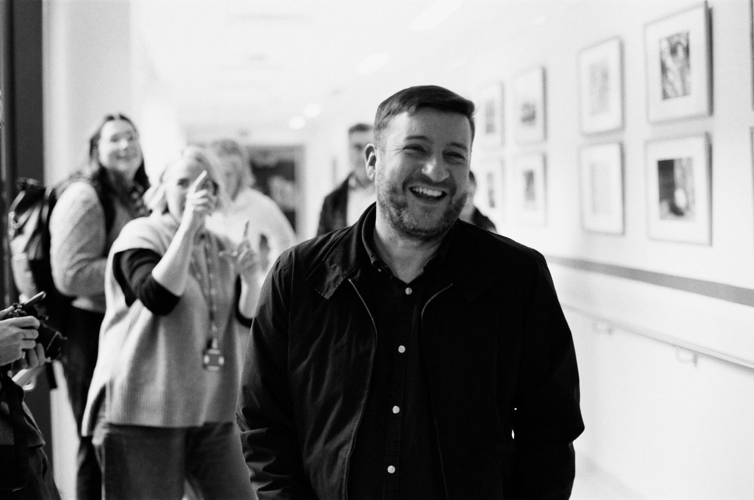 Man smiling in hallway with framed artwork on wall, black and white candid photo with people laughing in background.