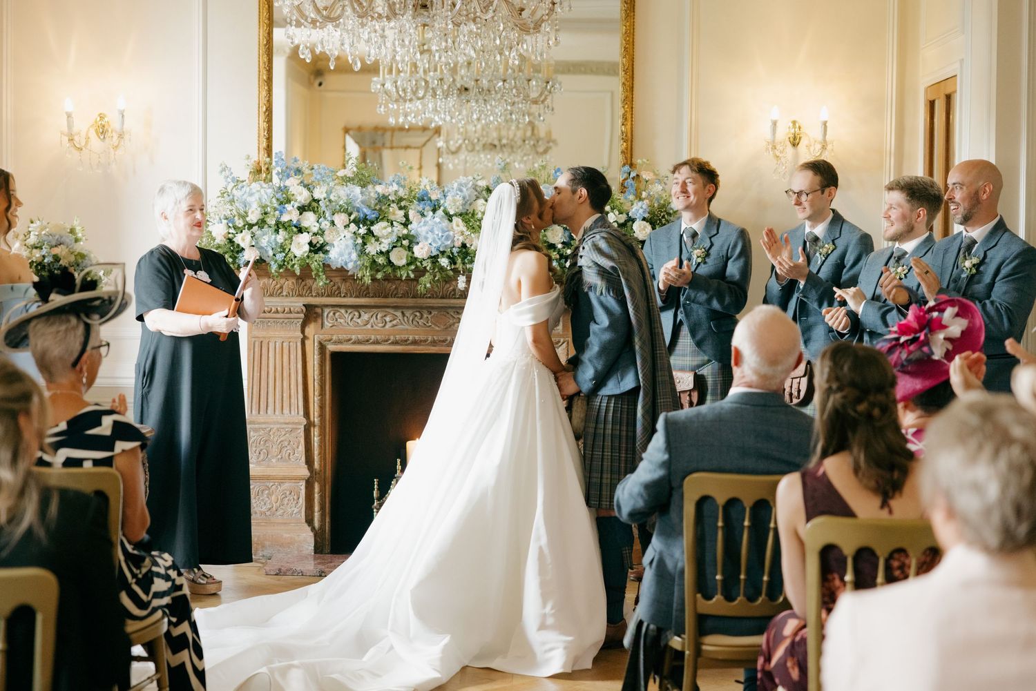 Bride and groom share first kiss at elegant indoor wedding ceremony with guests applauding.