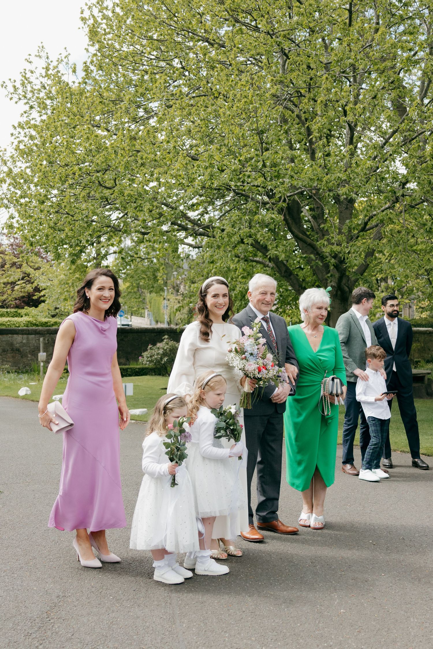 Wedding party walks outdoors, bride in white gown with family, flower girls, and guests amid lush green trees.