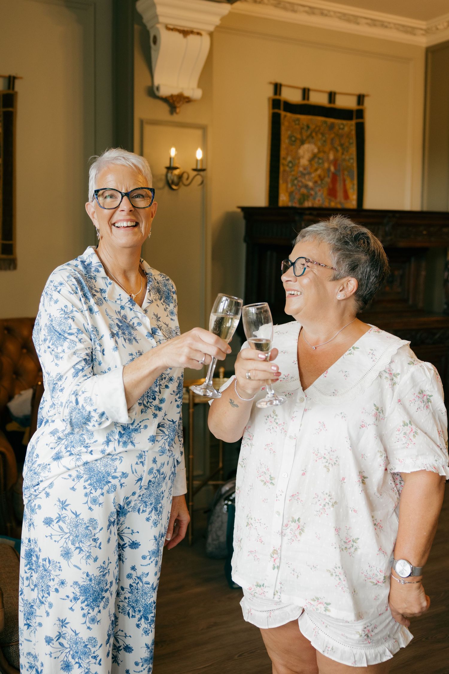 Two older women in floral pajamas toasting with champagne glasses, smiling in an elegant room.