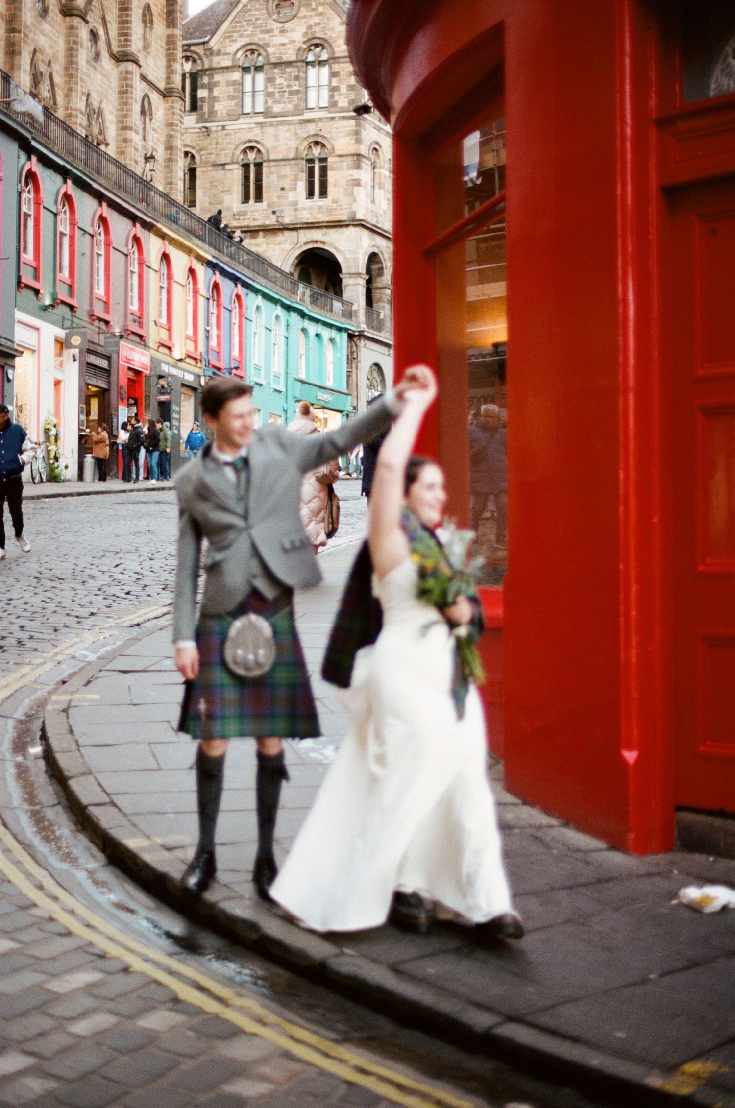 Joyful newlyweds walk Edinburgh's Victoria Street, groom in kilt, bride in white gown raising hand.