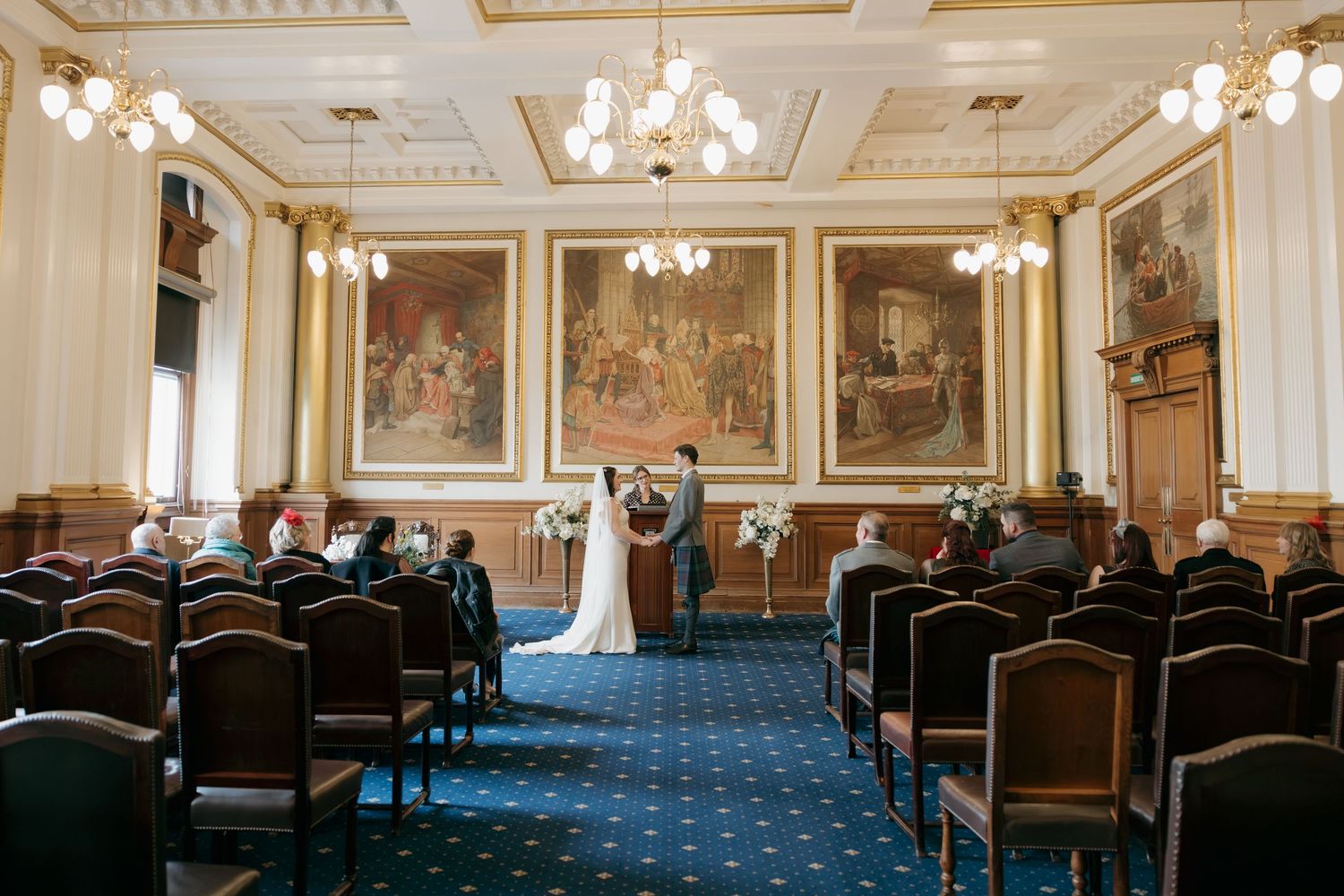 Elegant wedding ceremony in a grand hall with ornate paintings, blue carpet, and wooden chairs with guests seated.