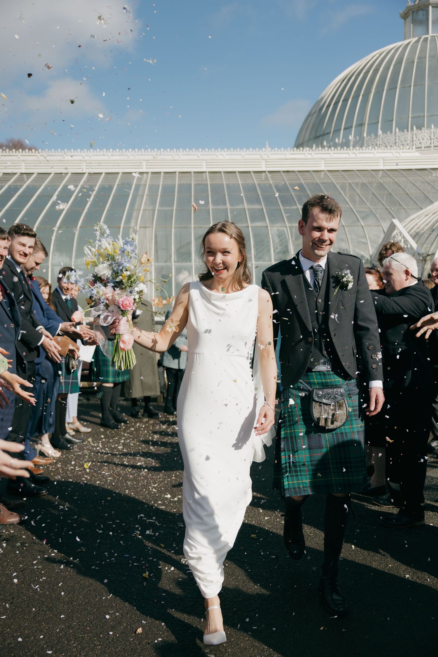 Bride in white dress and groom in tartan kilt walk through confetti outside a glass greenhouse wedding venue.