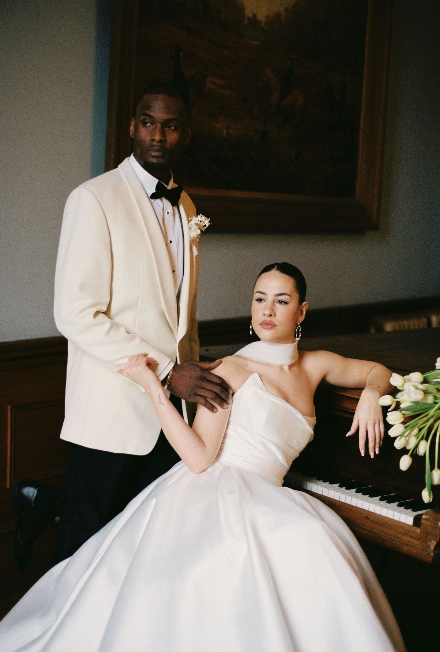 Elegant wedding couple posing by a piano, groom in ivory tuxedo and bride in strapless ball gown with white tulips.