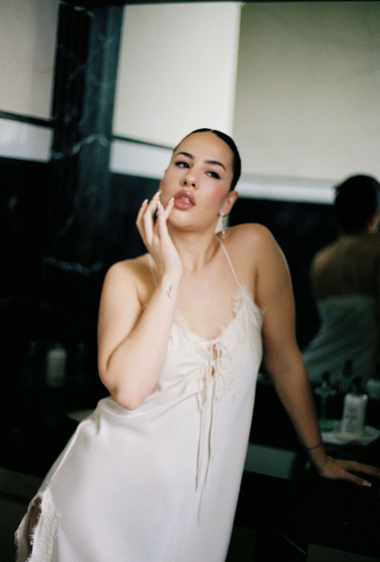 A woman wearing a white slip dress poses with her hand on her face, looking contemplative.