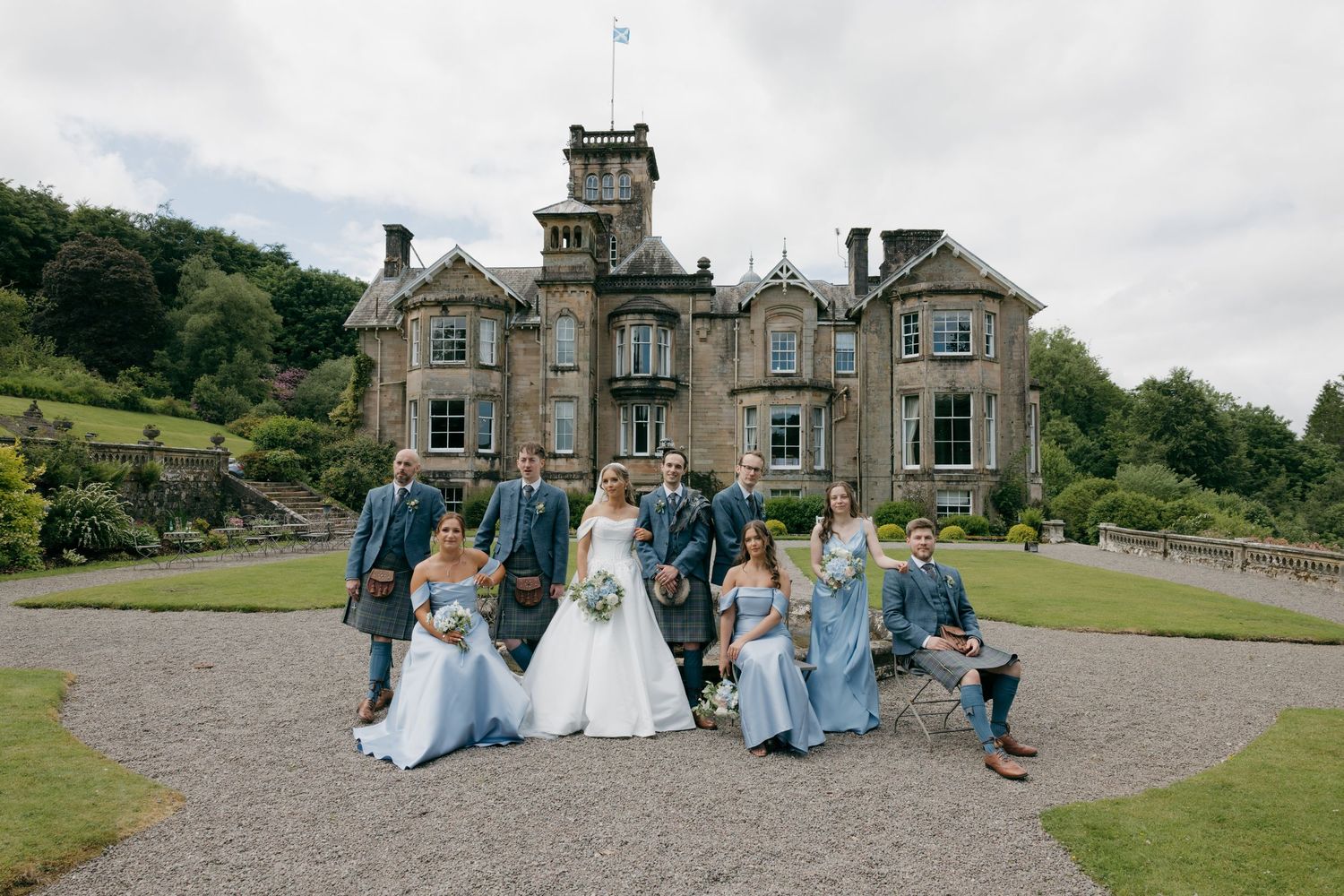 Wedding party poses in front of a grand Scottish castle with bridesmaids in blue dresses and groomsmen in kilts.