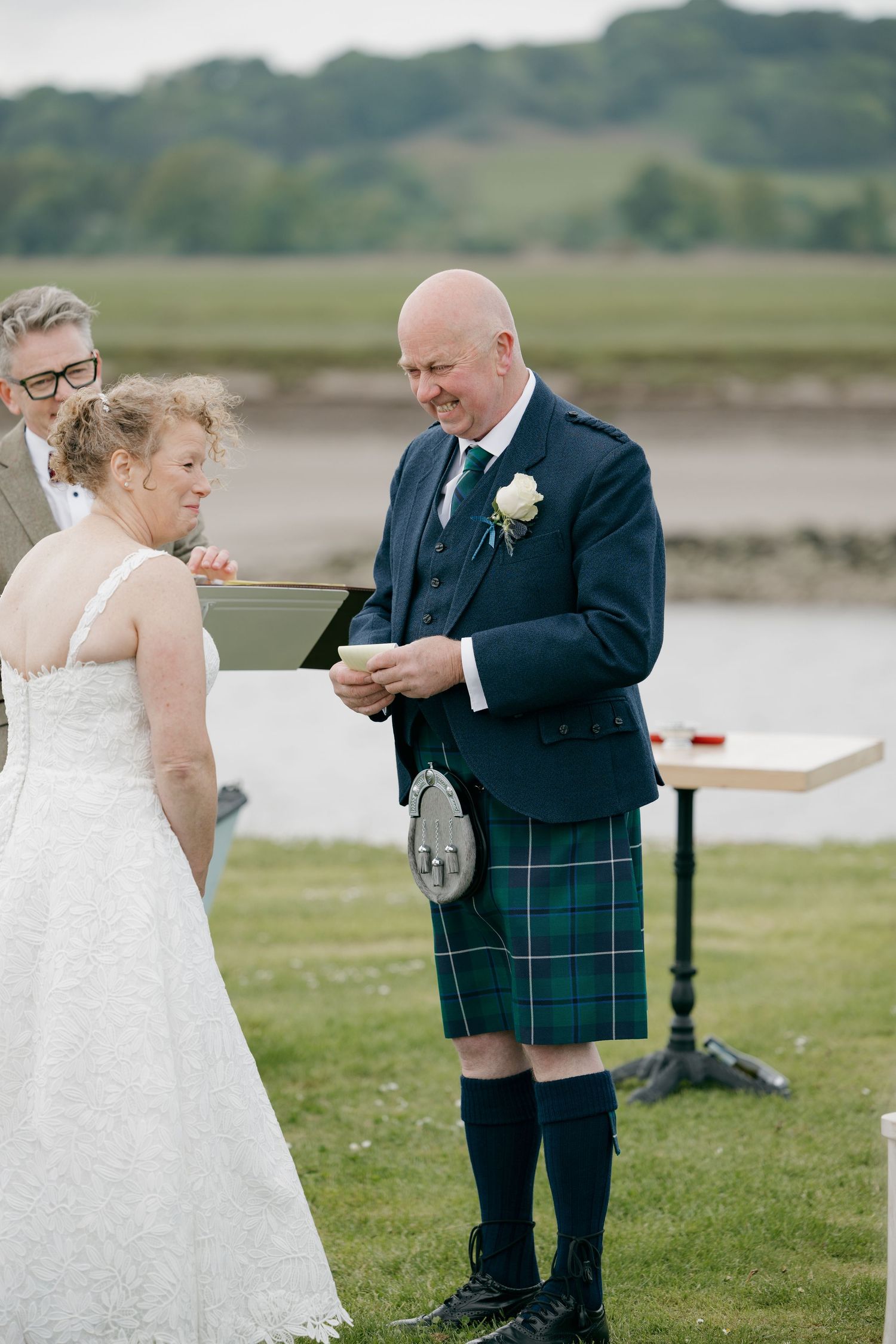 Groom in blue jacket and green tartan kilt smiling during outdoor Scottish wedding ceremony with bride in white lace gown.