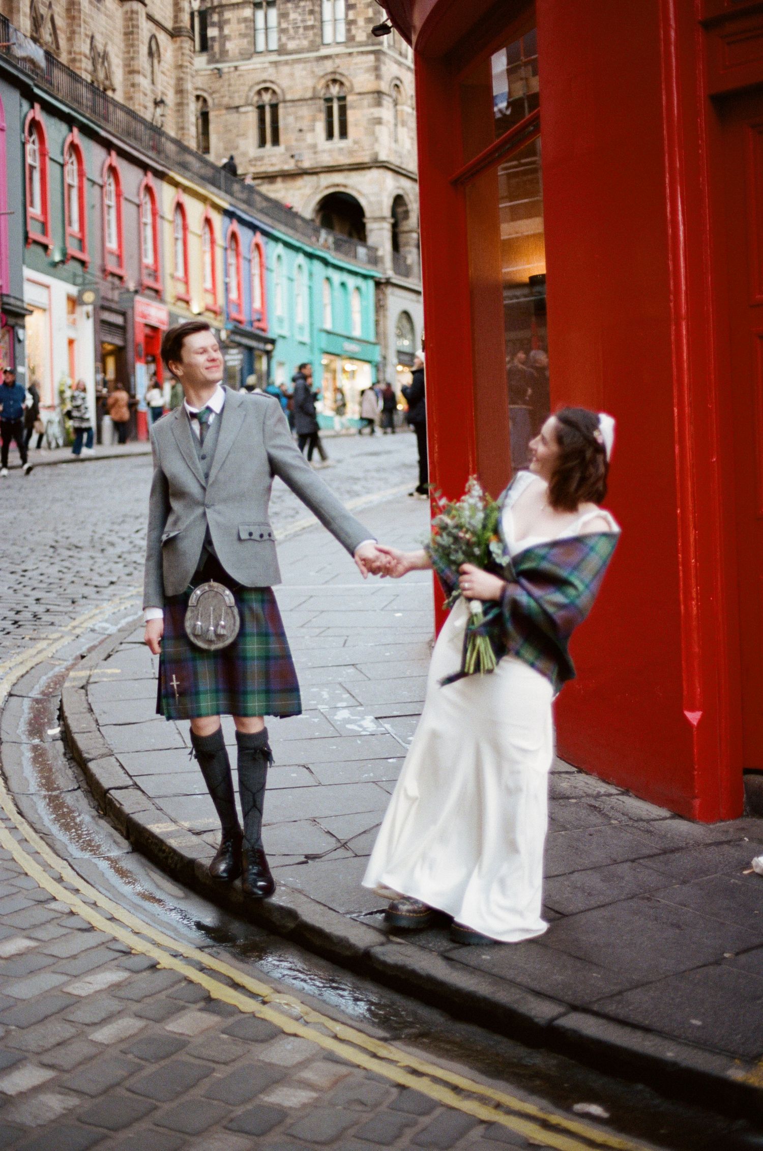 Newlyweds holding hands on a cobblestone street in Edinburgh, groom in tartan kilt, bride in white gown with plaid shawl.
