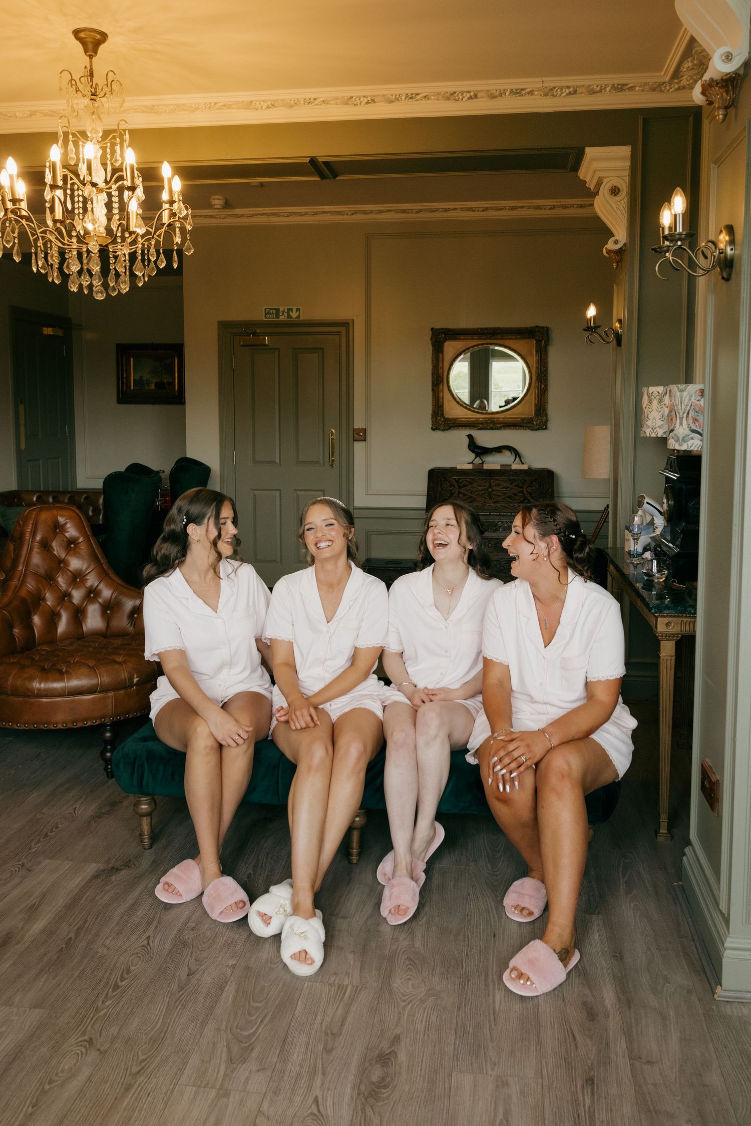 Four bridesmaids in white robes and pink slippers sitting together laughing in a luxurious room.