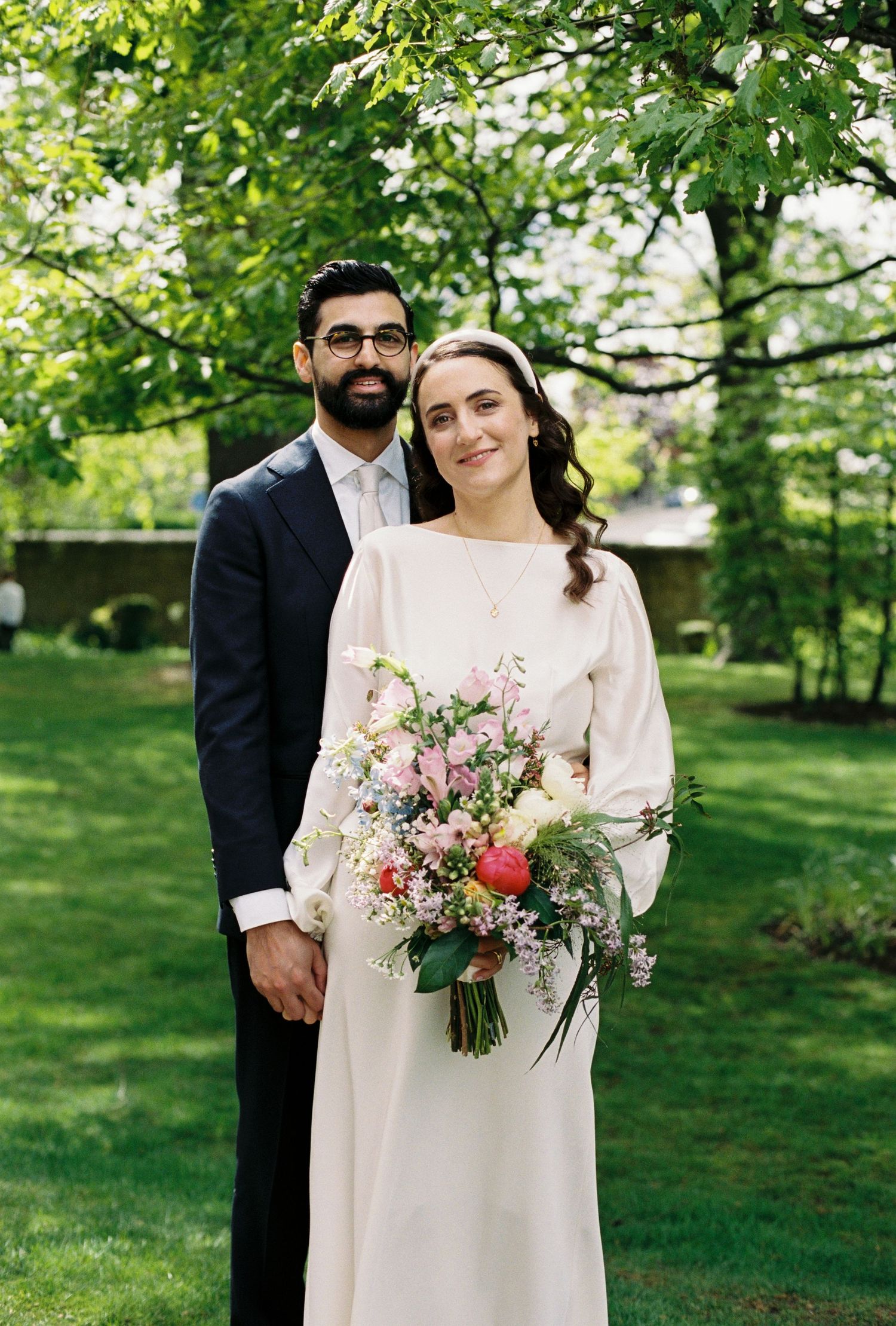 A couple poses outdoors on their wedding day, bride in white gown holding colorful bouquet, groom in navy suit.