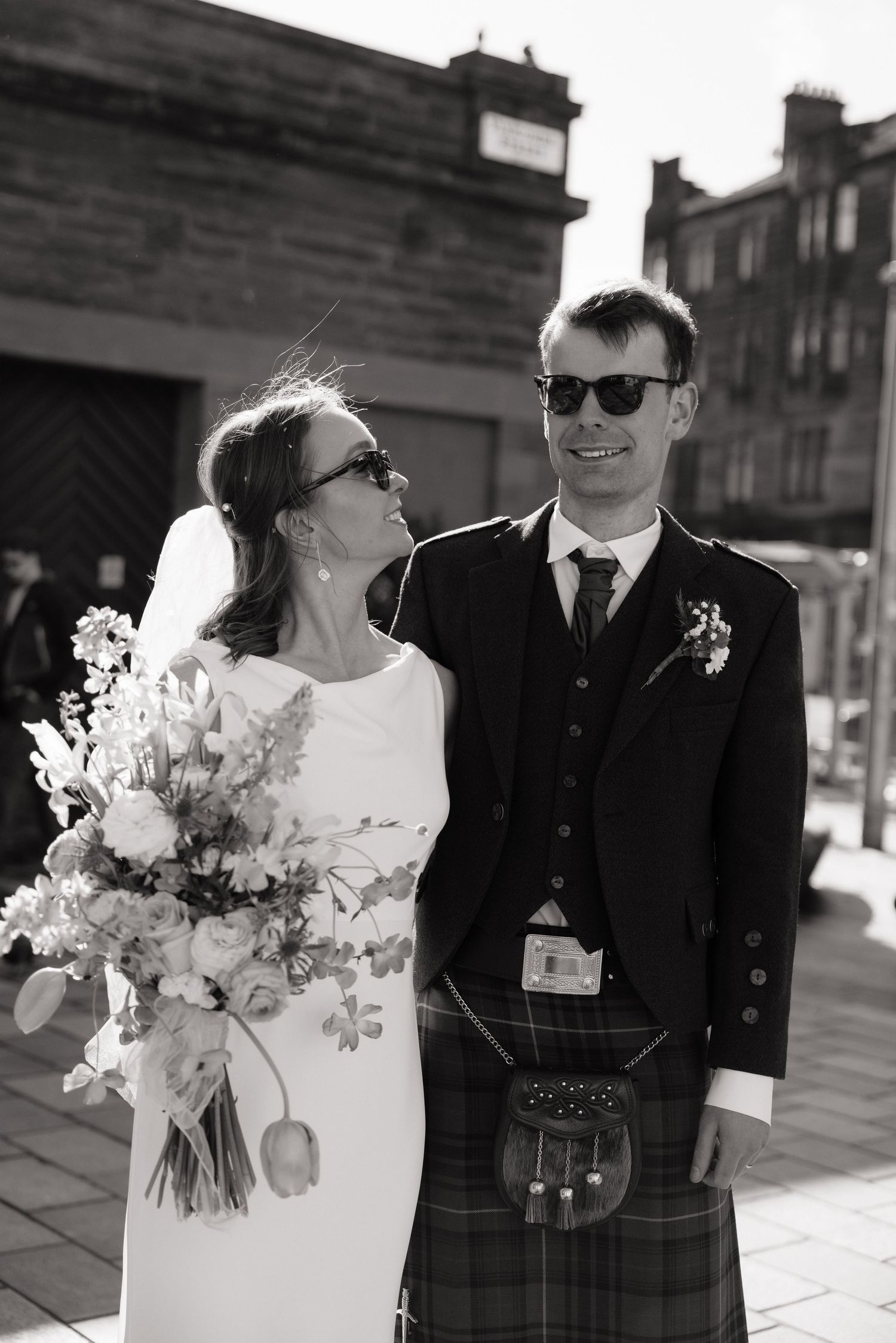 Black and white photo of a bride in white dress and groom in Scottish kilt posing outdoors on their wedding day.