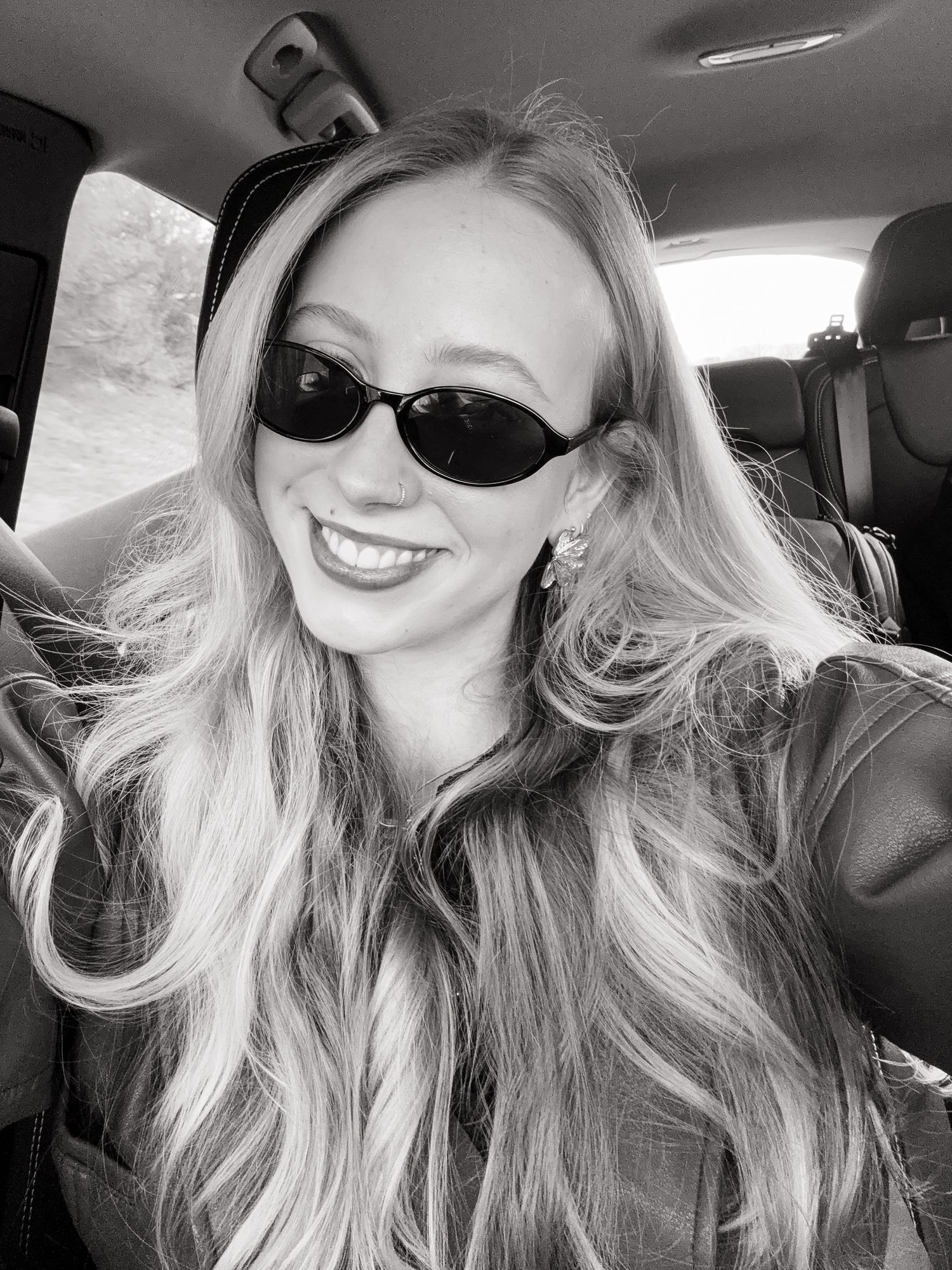 Smiling blonde woman wearing small oval sunglasses takes a selfie inside a car, black and white photo.
