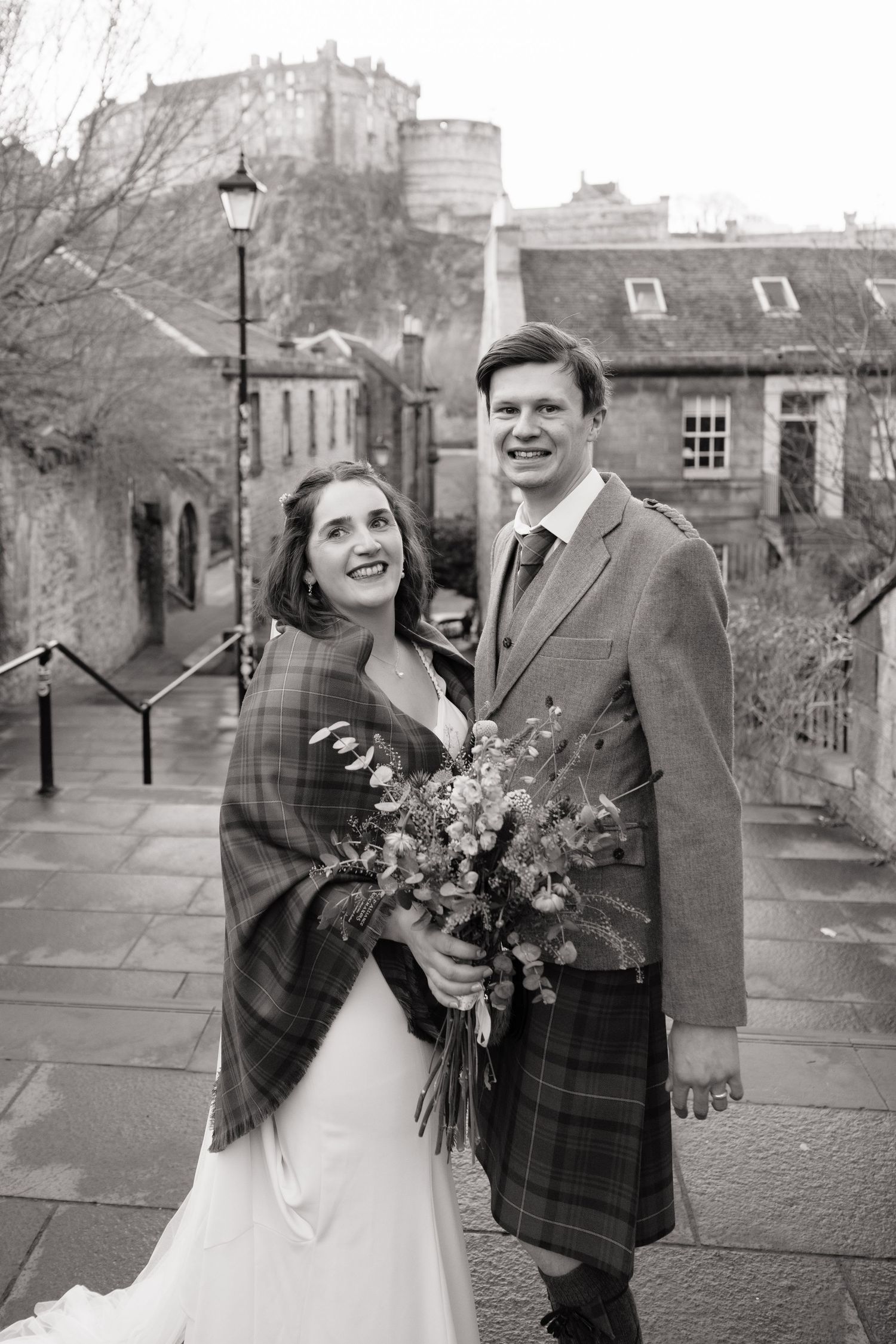 Black and white photo of a smiling bride and groom in a kilt posing with a bouquet near Edinburgh Castle.