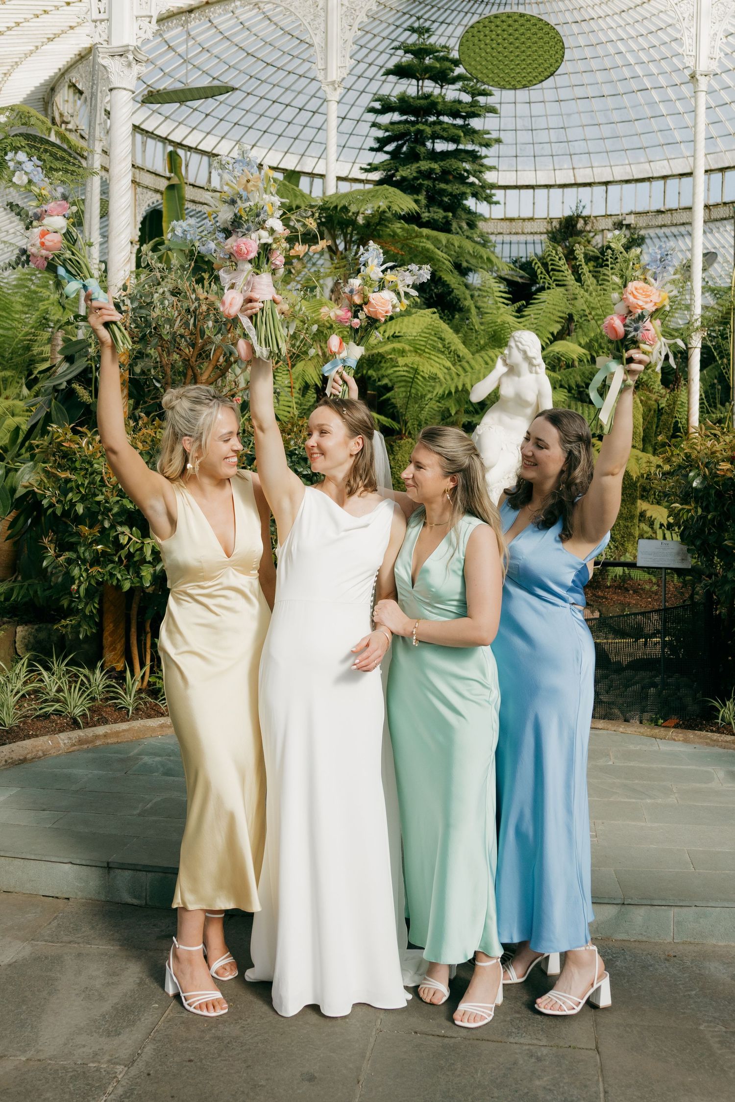 Bride in white gown with bridesmaids in pastel satin dresses raising floral bouquets in a lush greenhouse setting.
