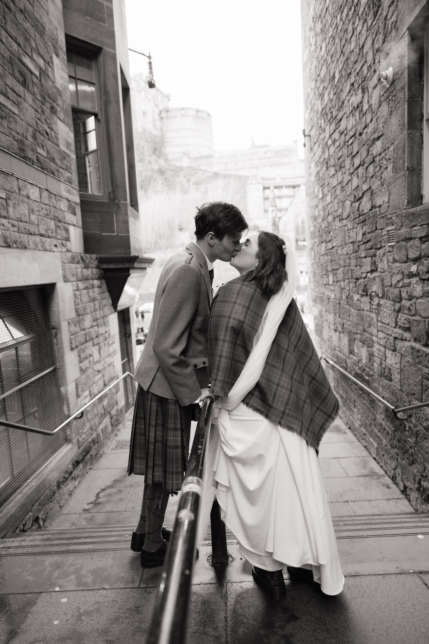 Couple kissing on stone steps in Edinburgh, groom in tartan kilt, bride in white dress with plaid wrap.