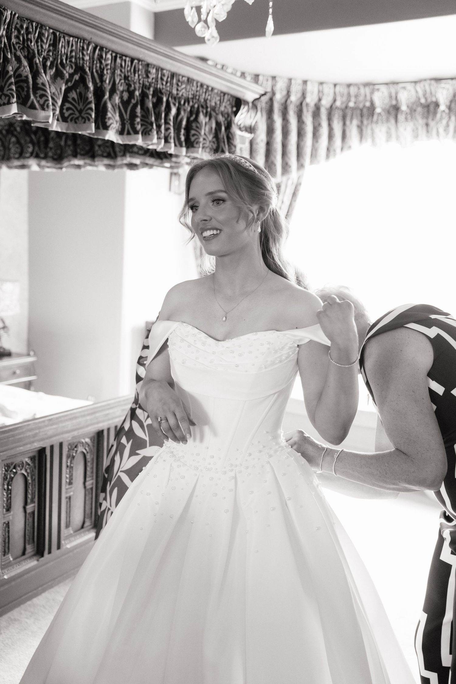 Black and white photo of smiling bride in off-shoulder ballgown being helped to dress indoors.