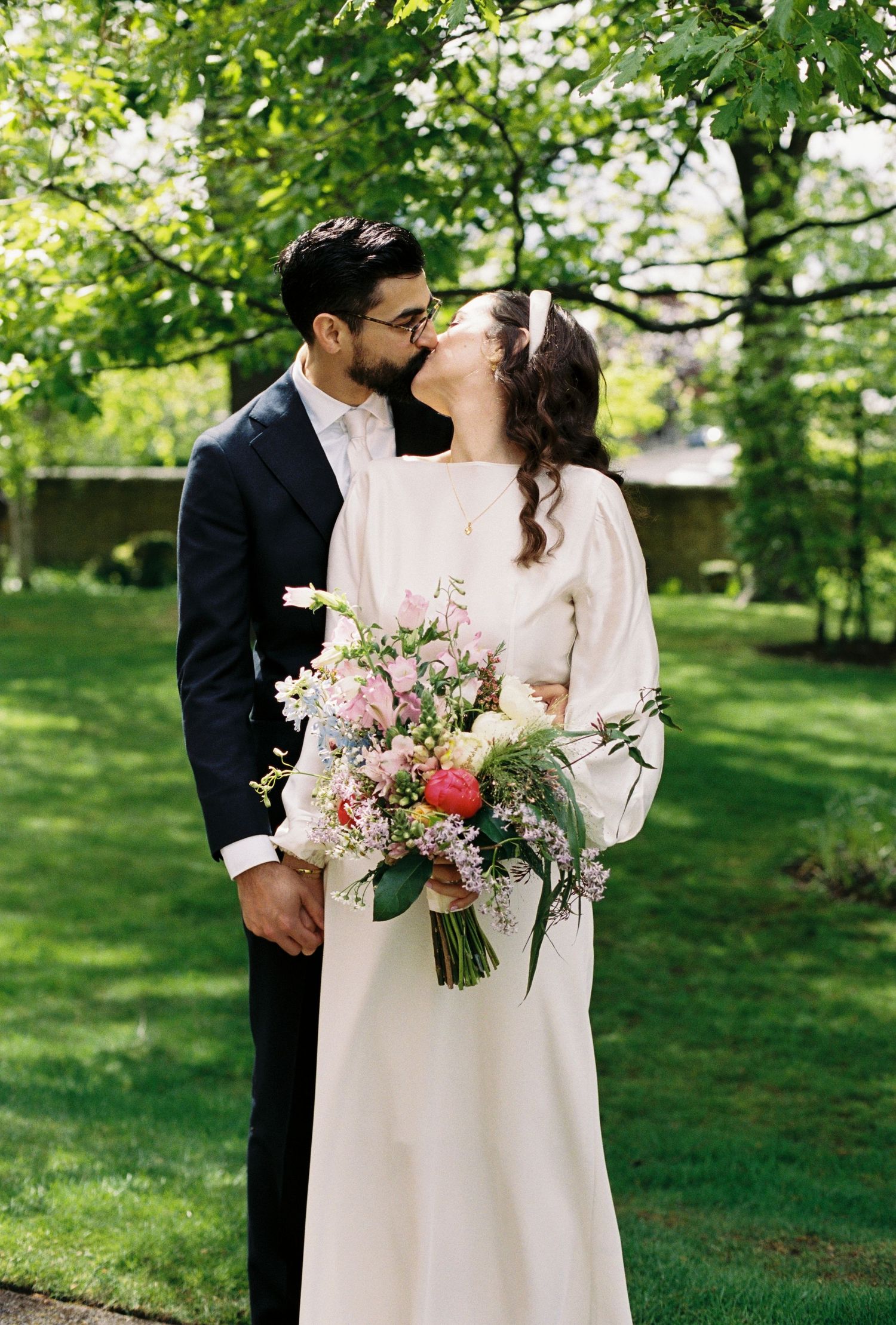Couple sharing a romantic kiss outdoors, bride holding a pink floral bouquet in a white gown, groom in a dark suit.
