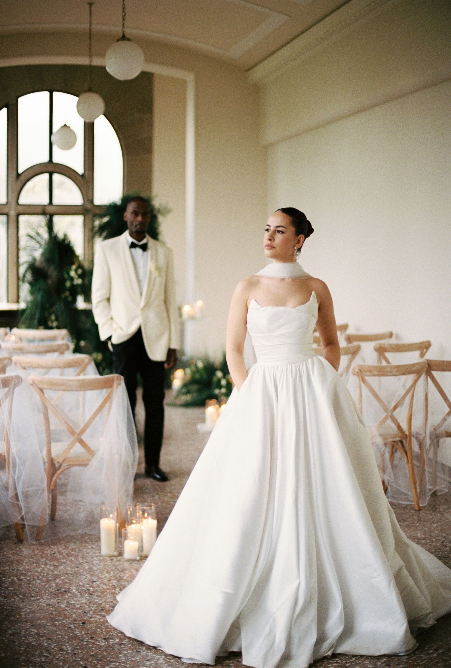A couple dressed in formal attire stands in a wedding venue, with the bride wearing a large white wedding dress.