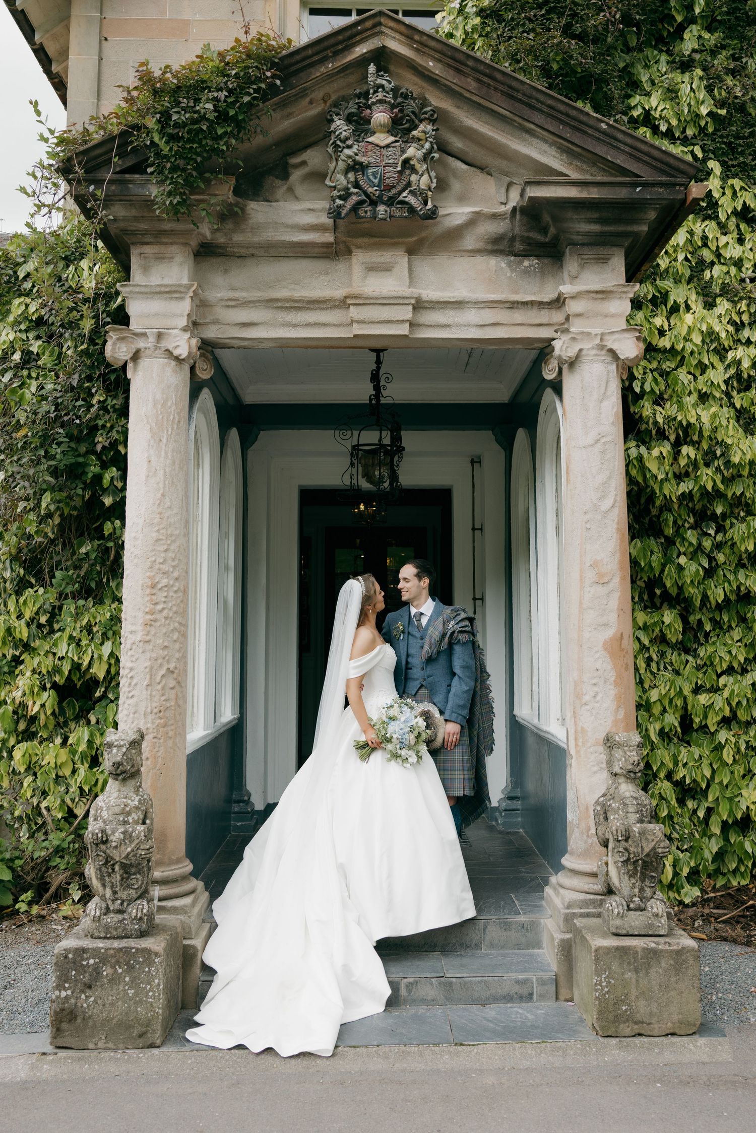 Bride and groom pose under an elegant stone portico with ivy-covered columns at a classic wedding venue.
