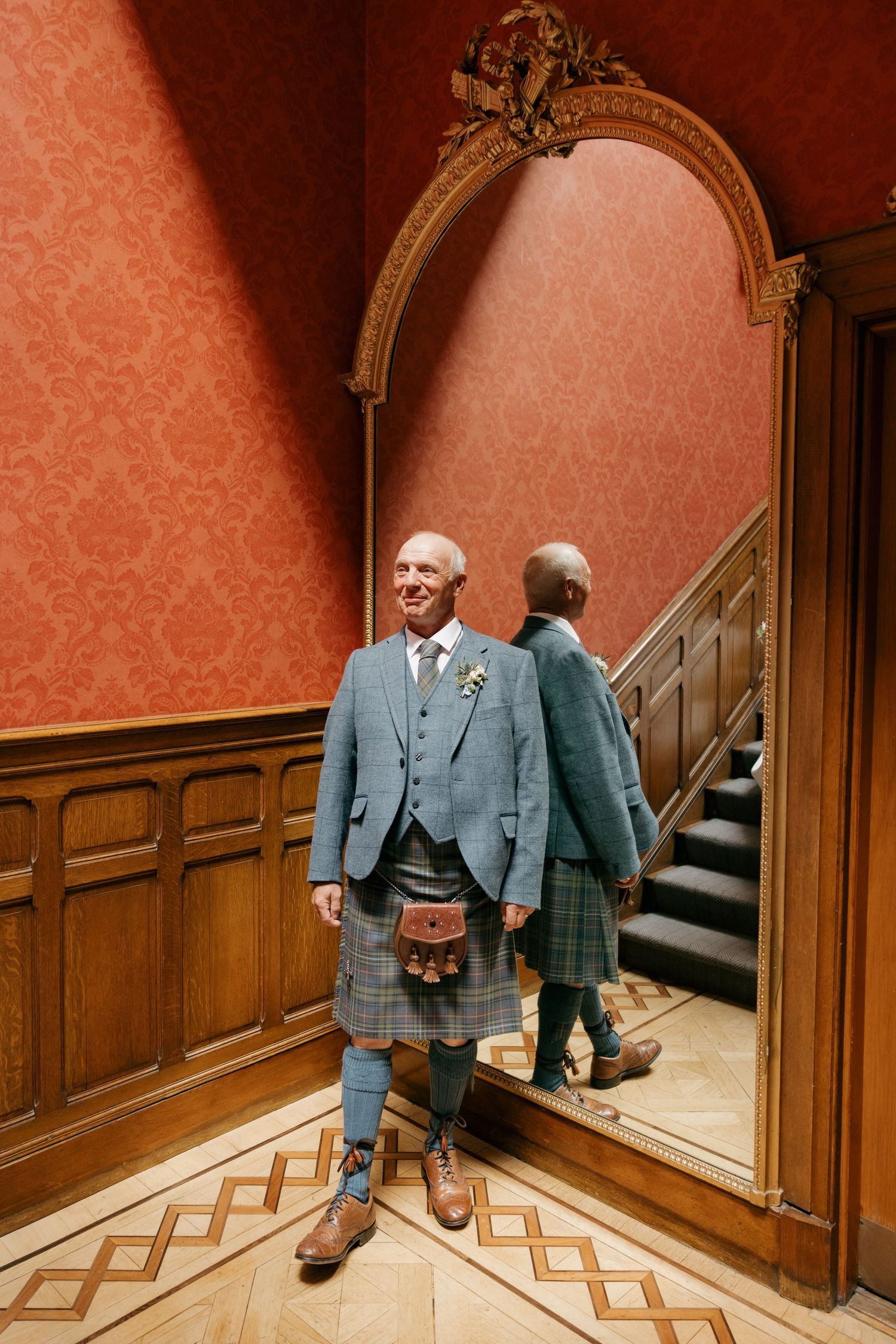 Elderly man in traditional Scottish kilt and tweed jacket stands before ornate mirror in elegant red-walled room.
