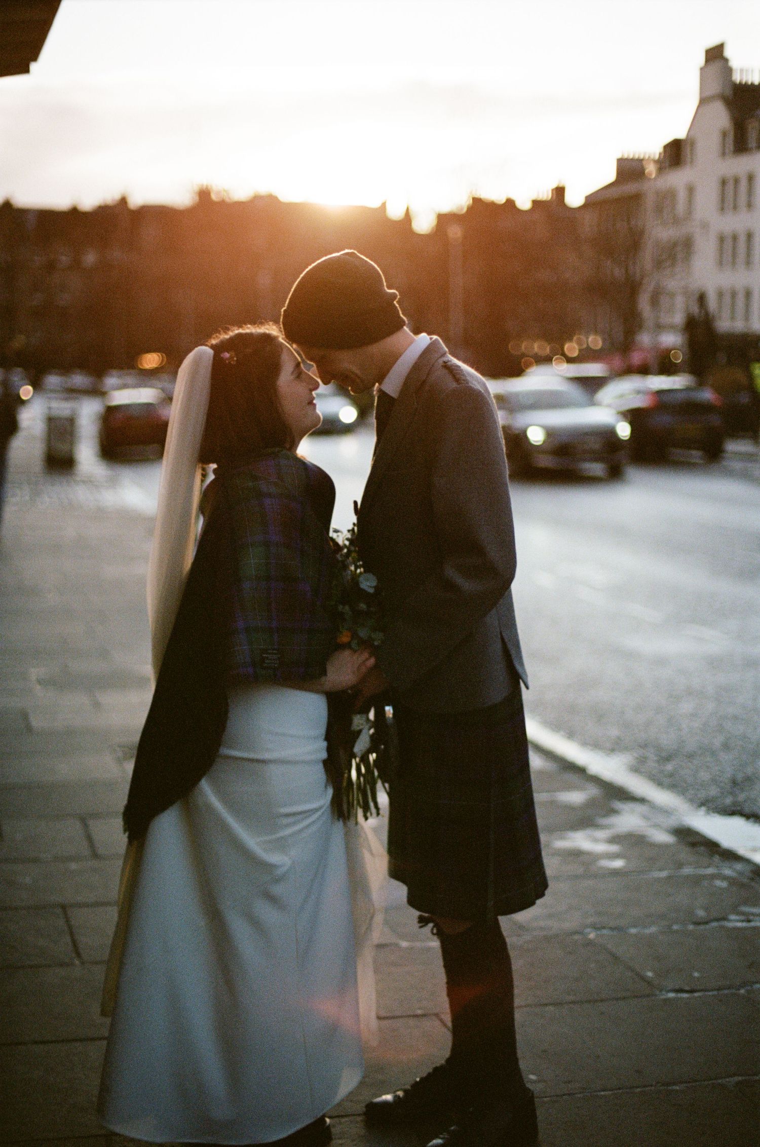 Wedding couple in tartan shawl and kilt share intimate moment on Edinburgh street bathed in golden sunset light.