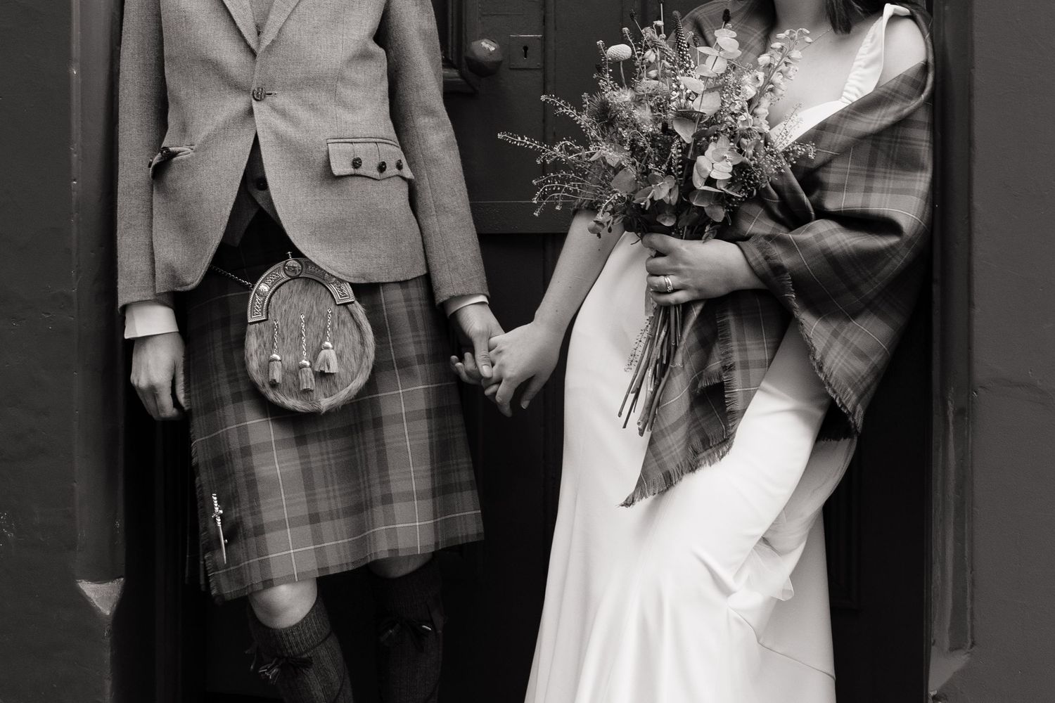 Black and white photo of Scottish wedding couple, groom in tartan kilt holding brides hand, bride holding wildflower bouquet.