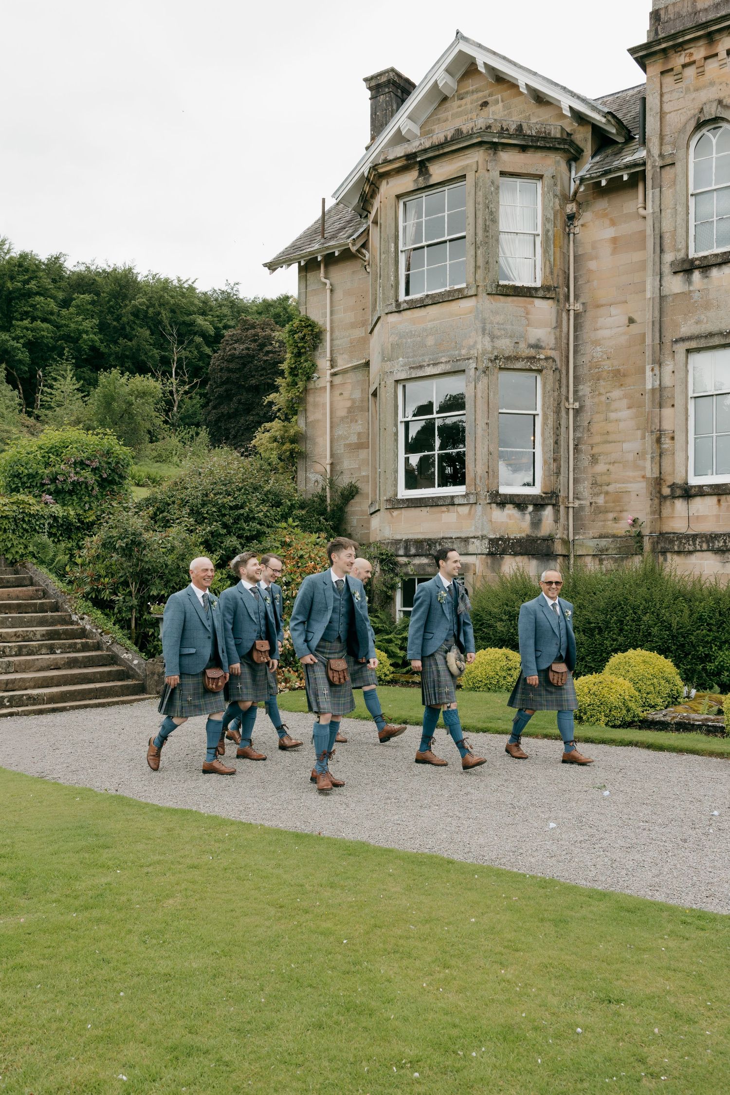 Groomsmen in matching blue tweed suits and tartan kilts walking outside a Scottish stone manor.