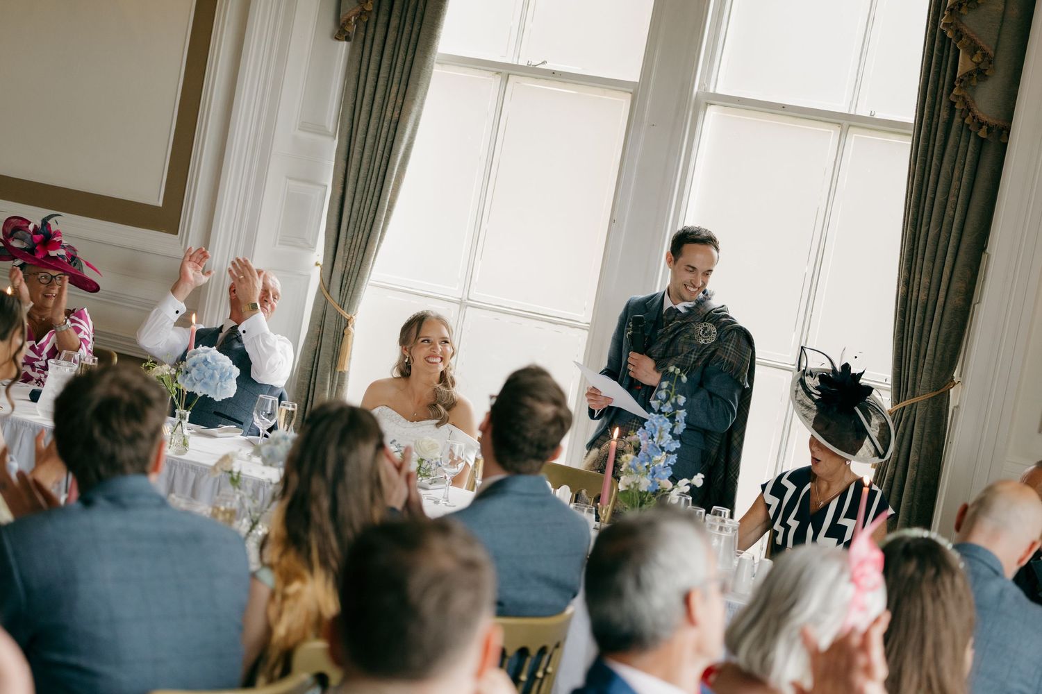 Groom in a kilt delivers a heartfelt speech to seated wedding guests in a bright elegant reception room.
