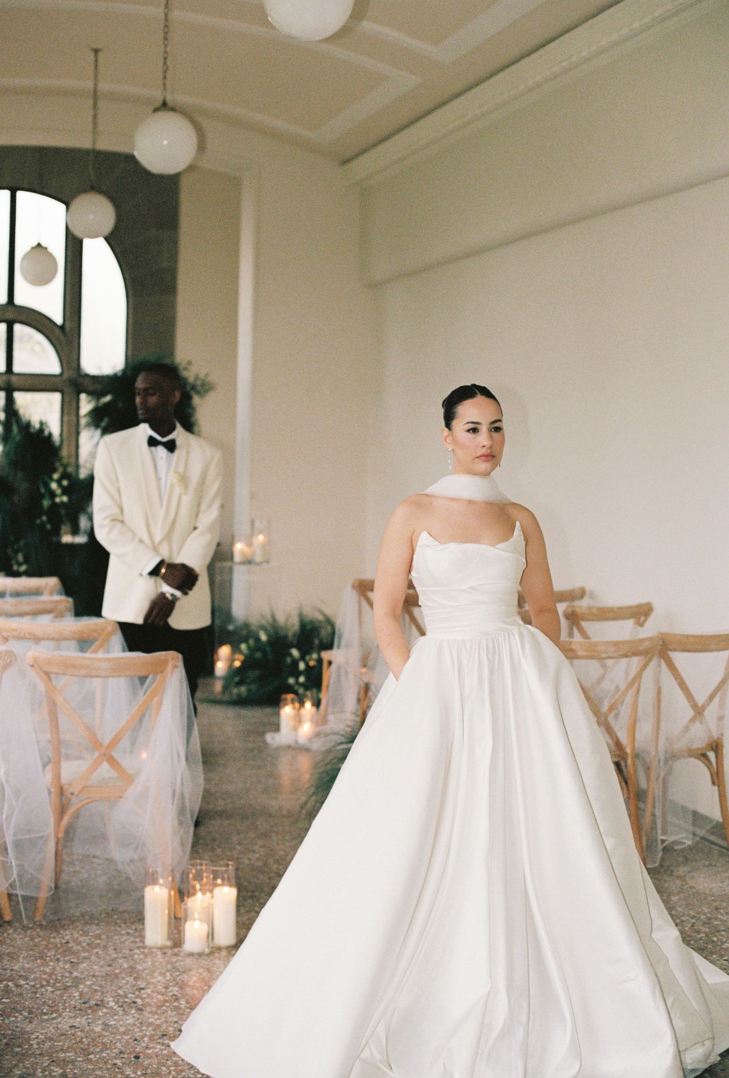 Bride in elegant white ball gown with choker neckline stands in candlelit ceremony venue as groom waits in white tuxedo.