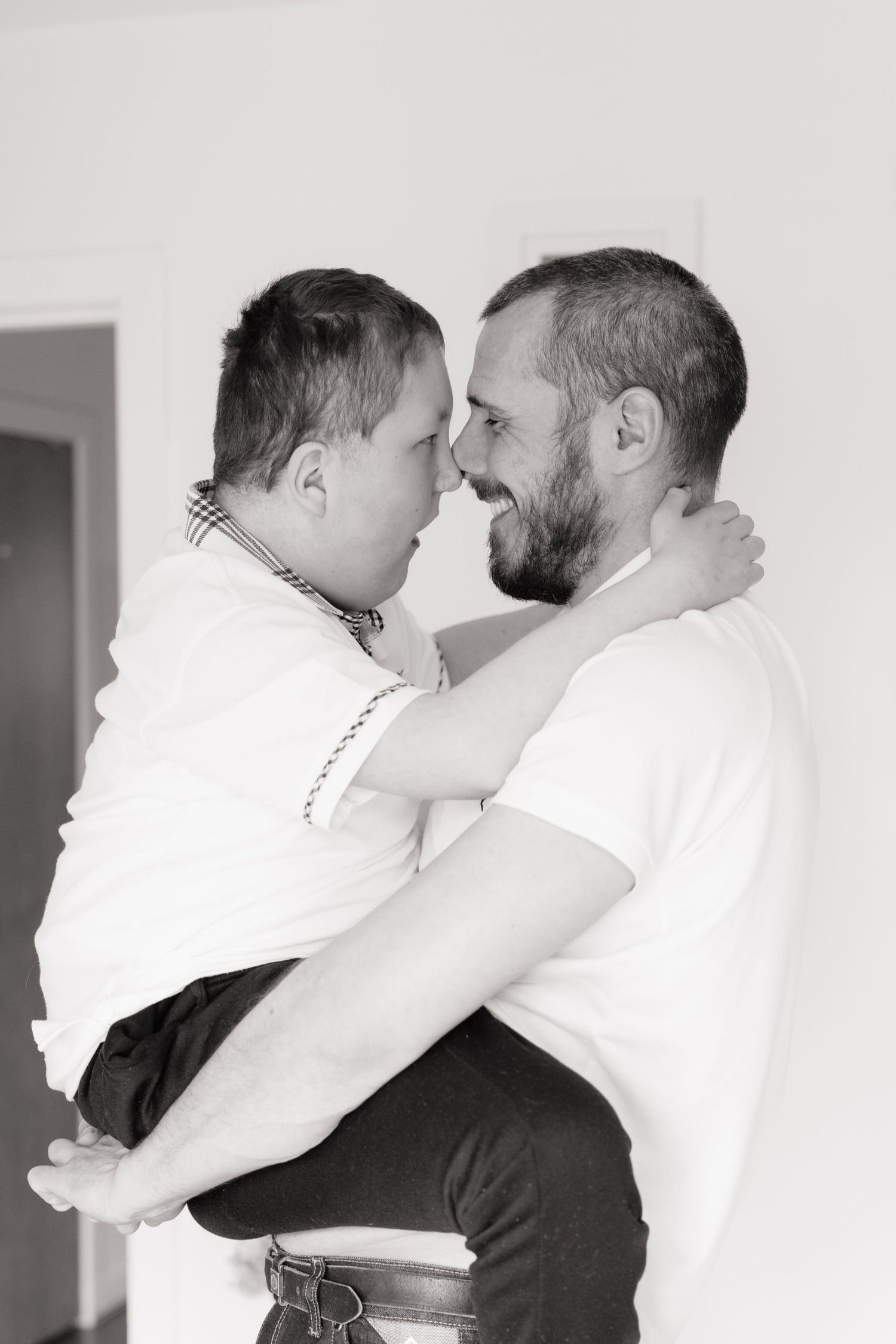 Black and white photo of a bearded man holding another man in his arms, sharing a joyful embrace indoors.