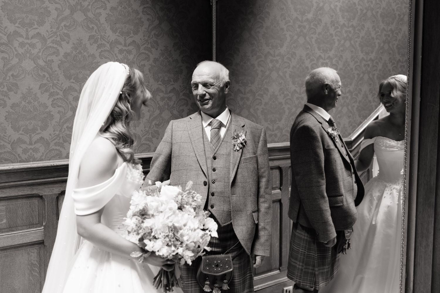 Bride in white gown with veil holds bouquet beside smiling older man in Scottish kilt at wedding venue.