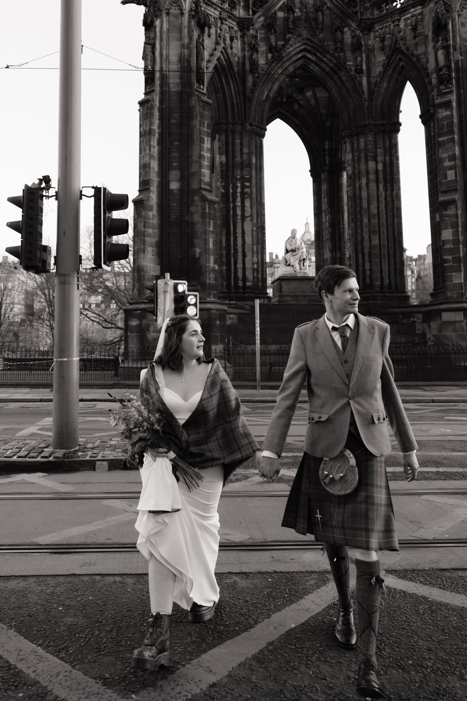 Newlyweds walk hand in hand past Scott Monument in Edinburgh, groom in tartan kilt, bride holding bouquet.