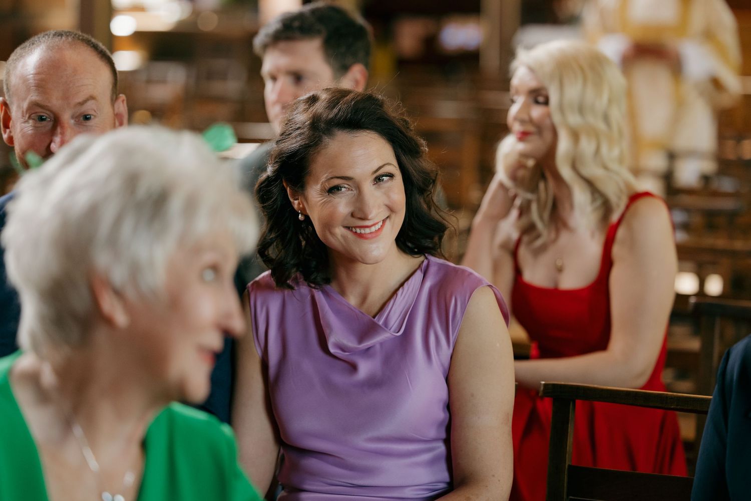 Women in colorful dresses smile and chat at an indoor social gathering in a warmly lit venue.