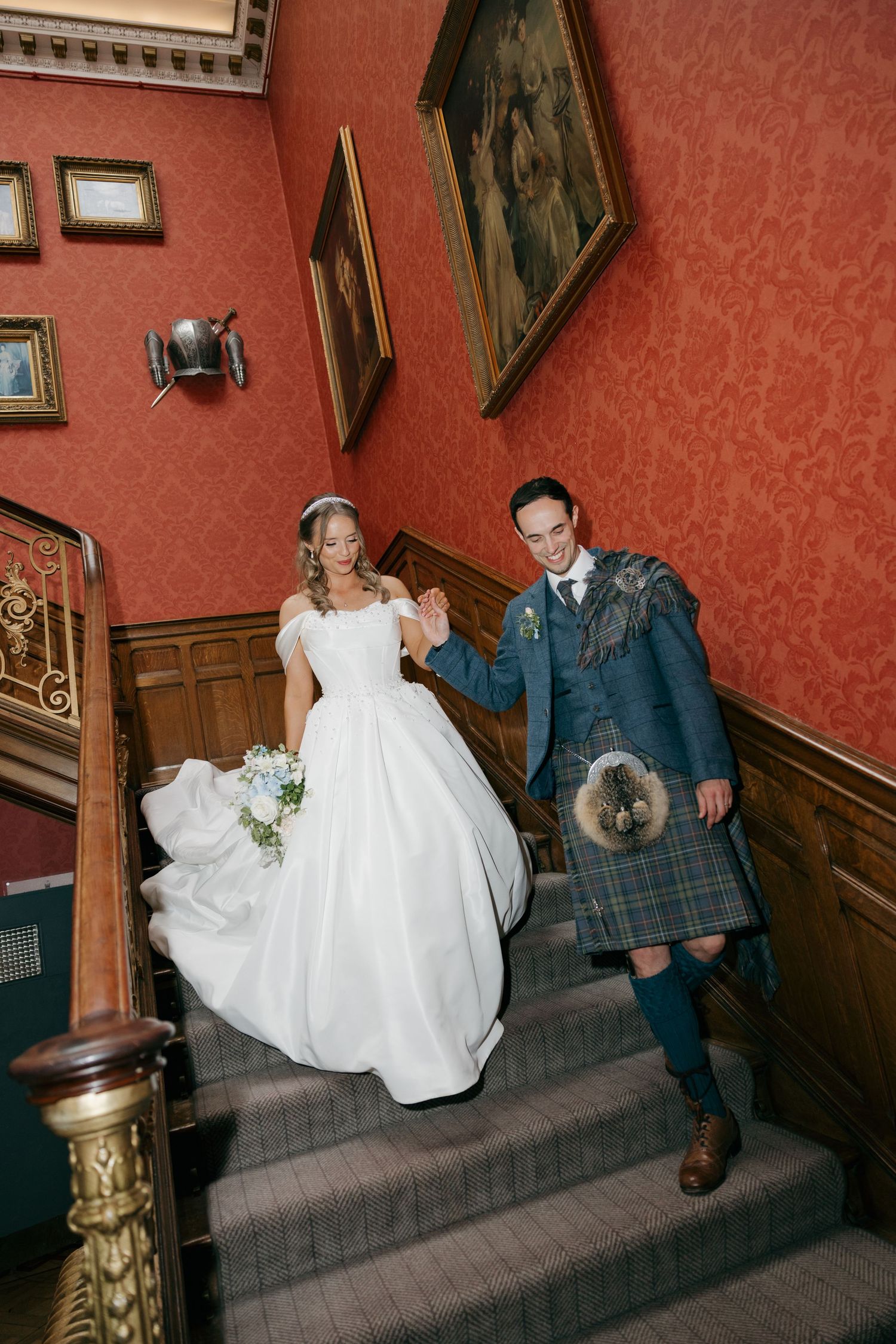 Smiling bride and groom in Scottish tartan kilt descend grand staircase in elegant red-walled manor.