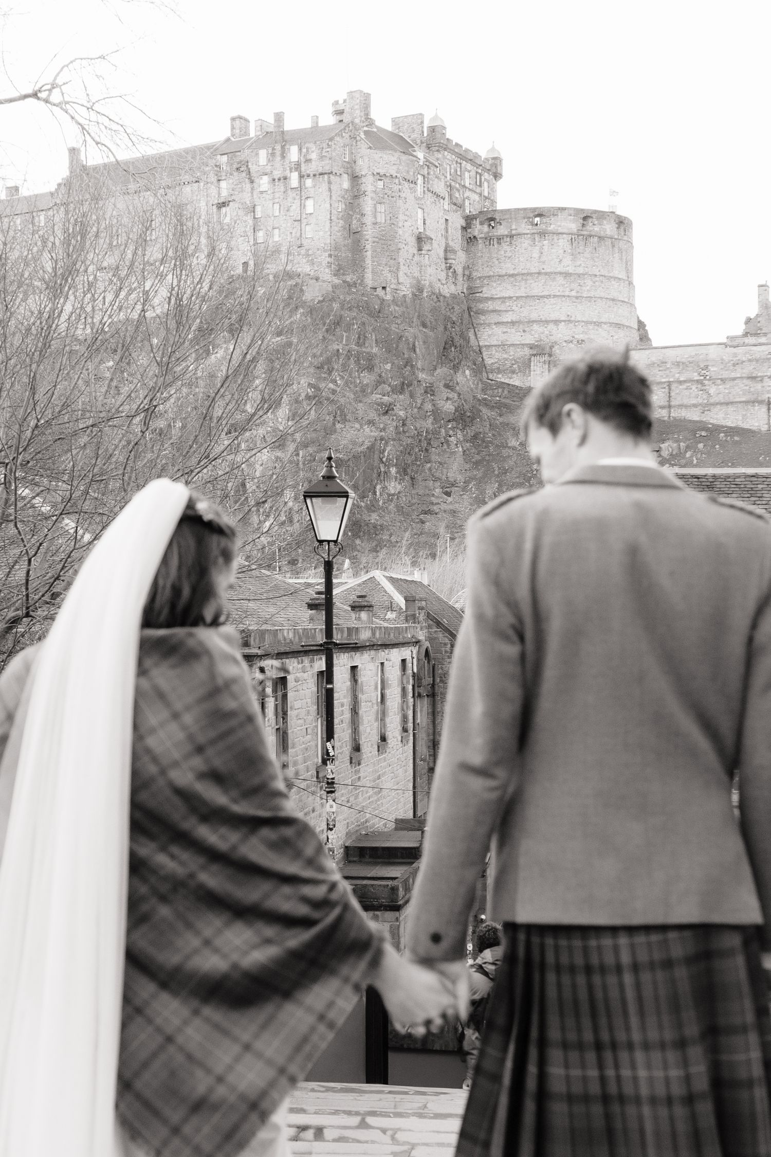 Black and white photo of a bride and groom in tartan holding hands facing Edinburgh Castle.