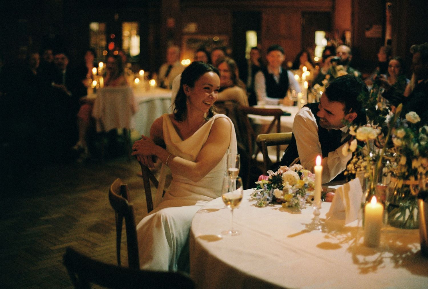 Bride in white dress sits at candlelit wedding reception table, sharing intimate moment with suited groom.