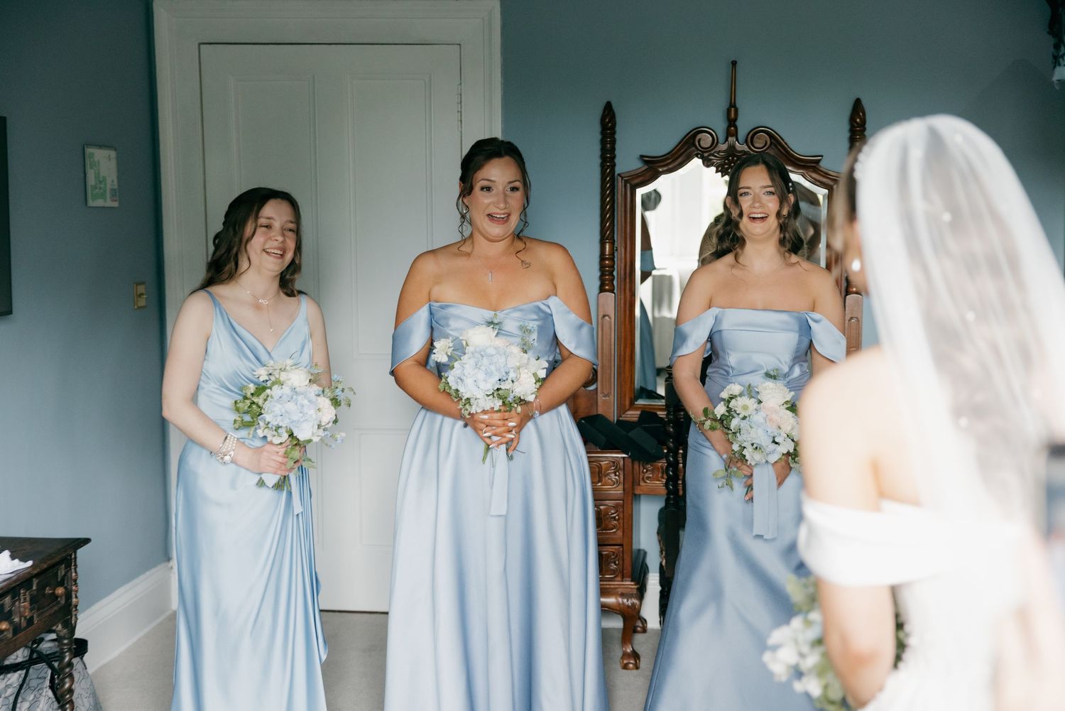 Three bridesmaids in light blue off-shoulder gowns holding white bouquets react to seeing the bride in a white wedding dress.