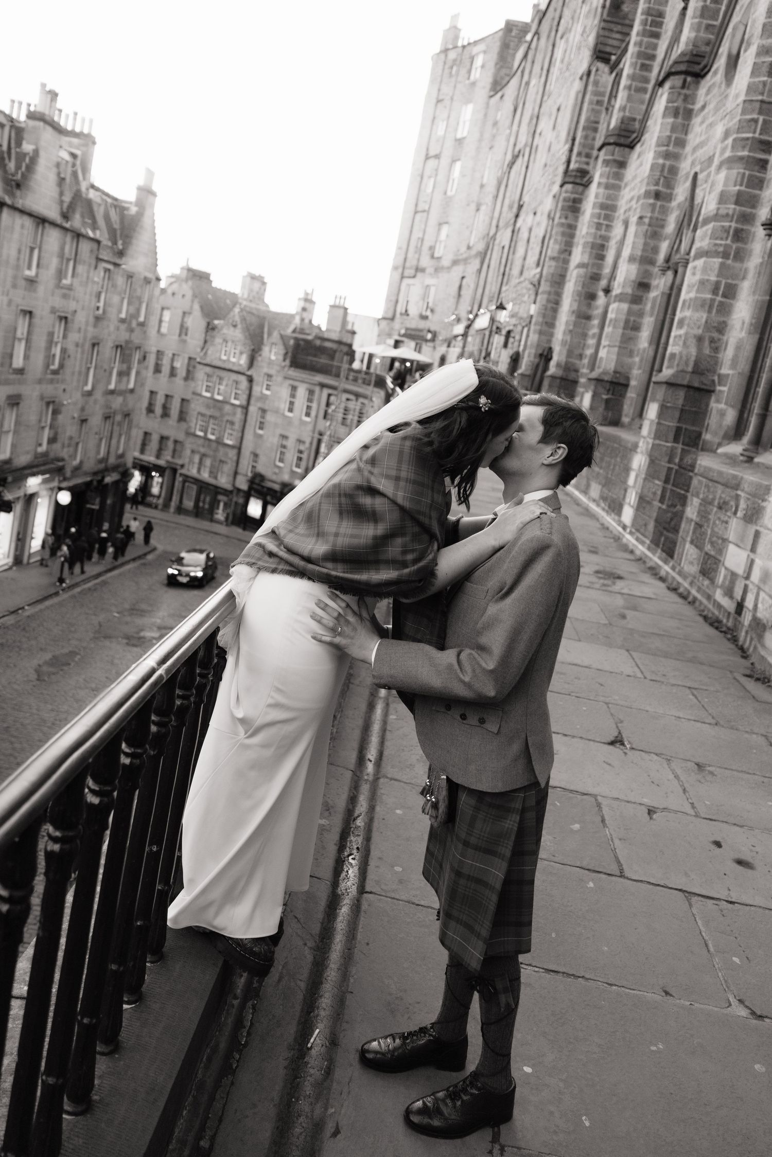 Bride and groom share romantic kiss on Edinburgh street balcony, groom wearing traditional Scottish kilt.