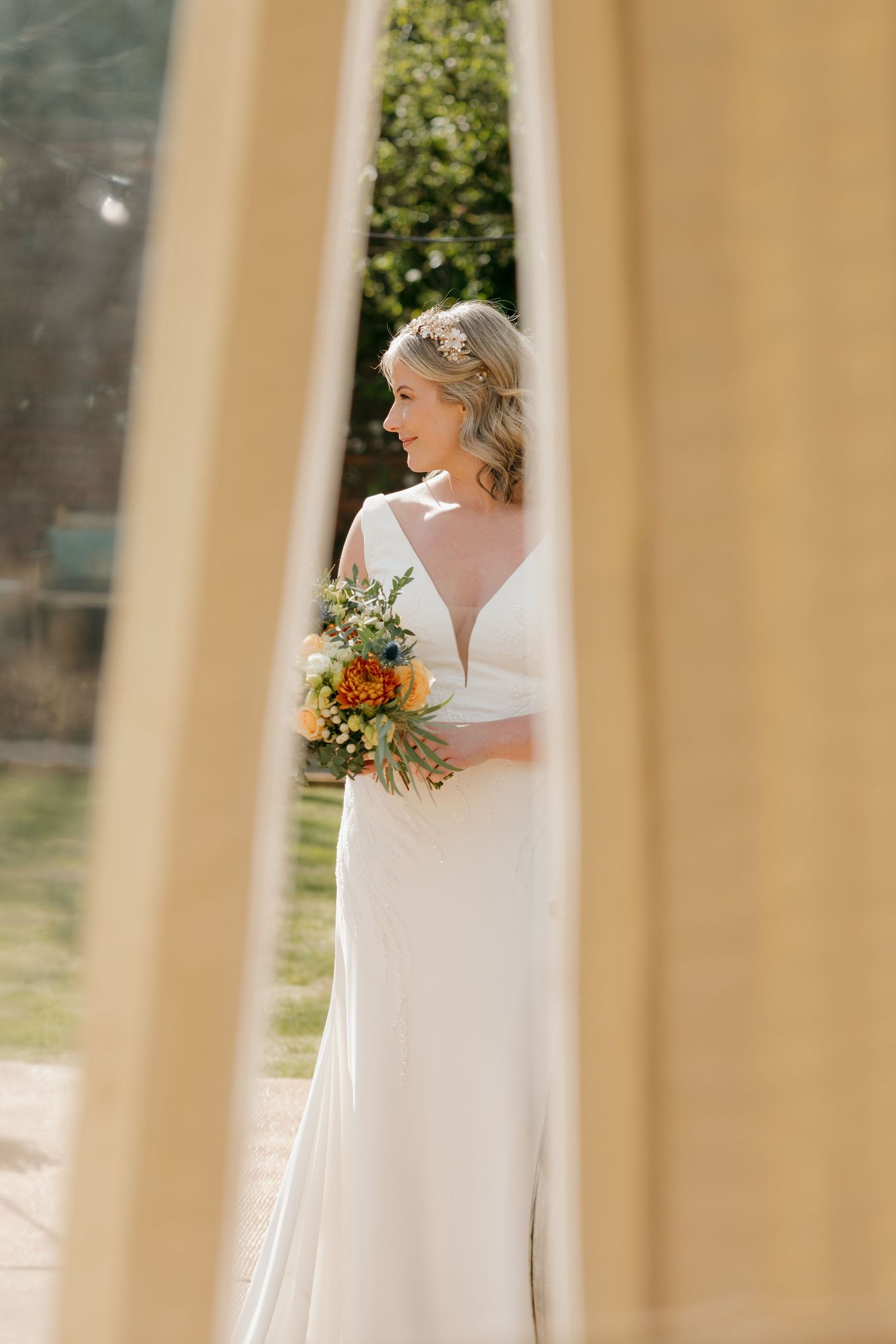 Bride in white gown holding colorful bouquet, viewed through doorframe in sunny outdoor setting.
