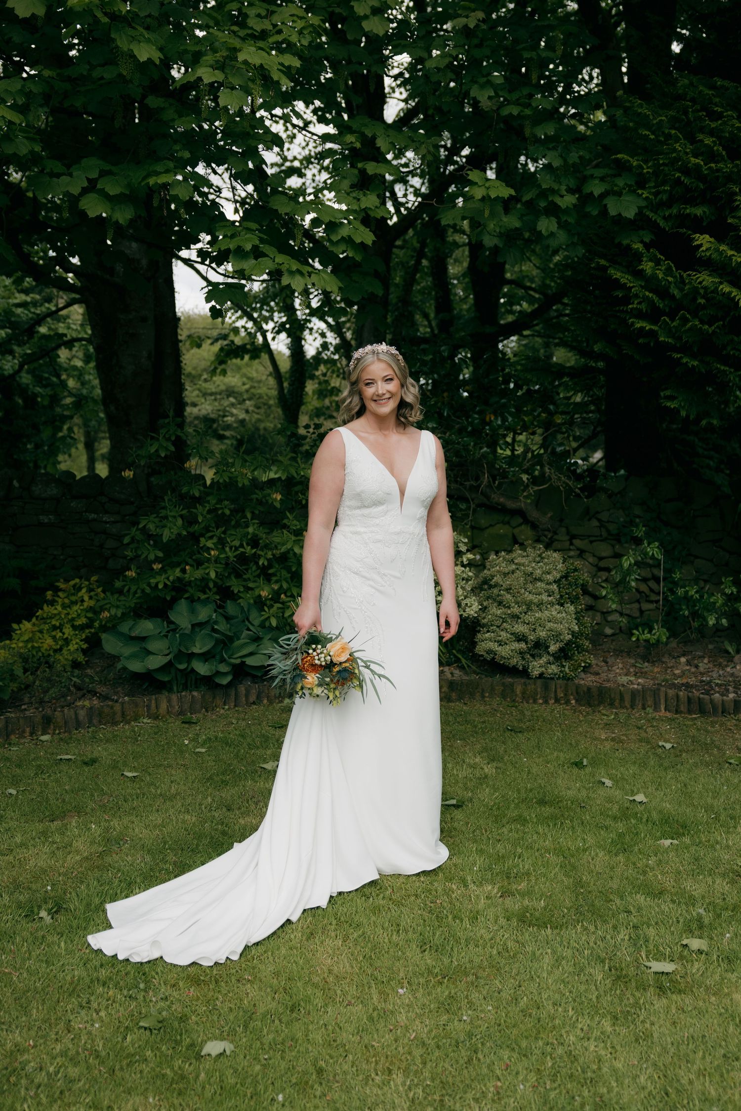 Bride in white V-neck wedding gown with train holds bouquet, standing on green lawn surrounded by lush trees.