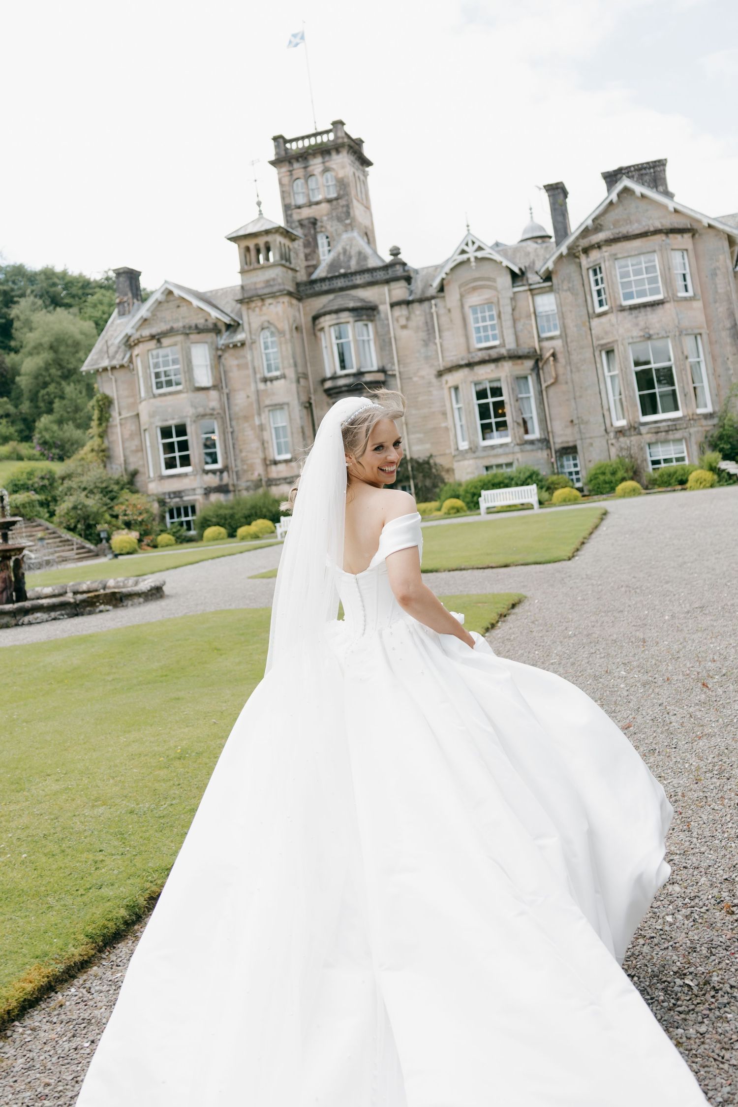 Elegant bride in off-shoulder gown with flowing train walks toward a Scottish country house estate.
