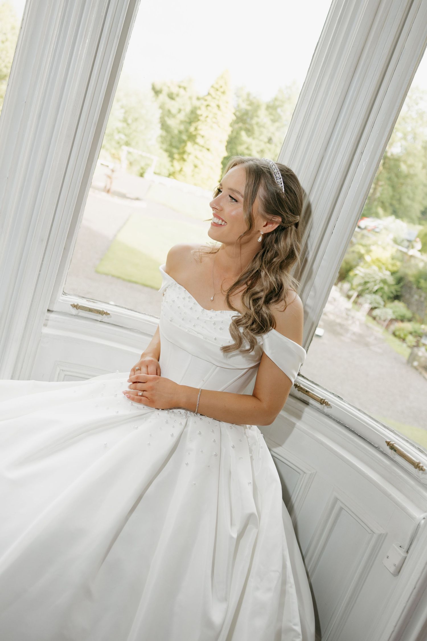 Radiant bride in off-shoulder white ballgown smiles while seated by large window in bright room.