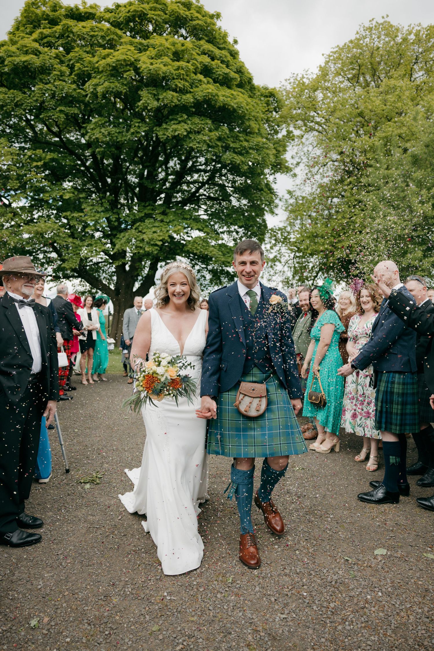 Bride and groom walk through confetti at Scottish wedding, groom wearing green tartan kilt, bride holding wildflower bouquet.