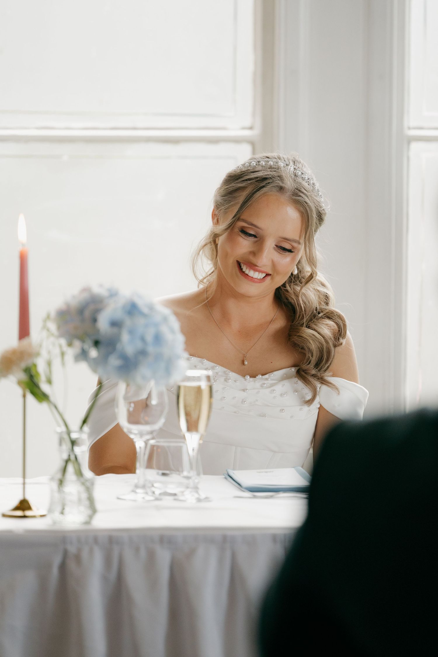 Smiling bride wearing a tiara and white off-shoulder gown sits at reception table with champagne and blue hydrangeas.