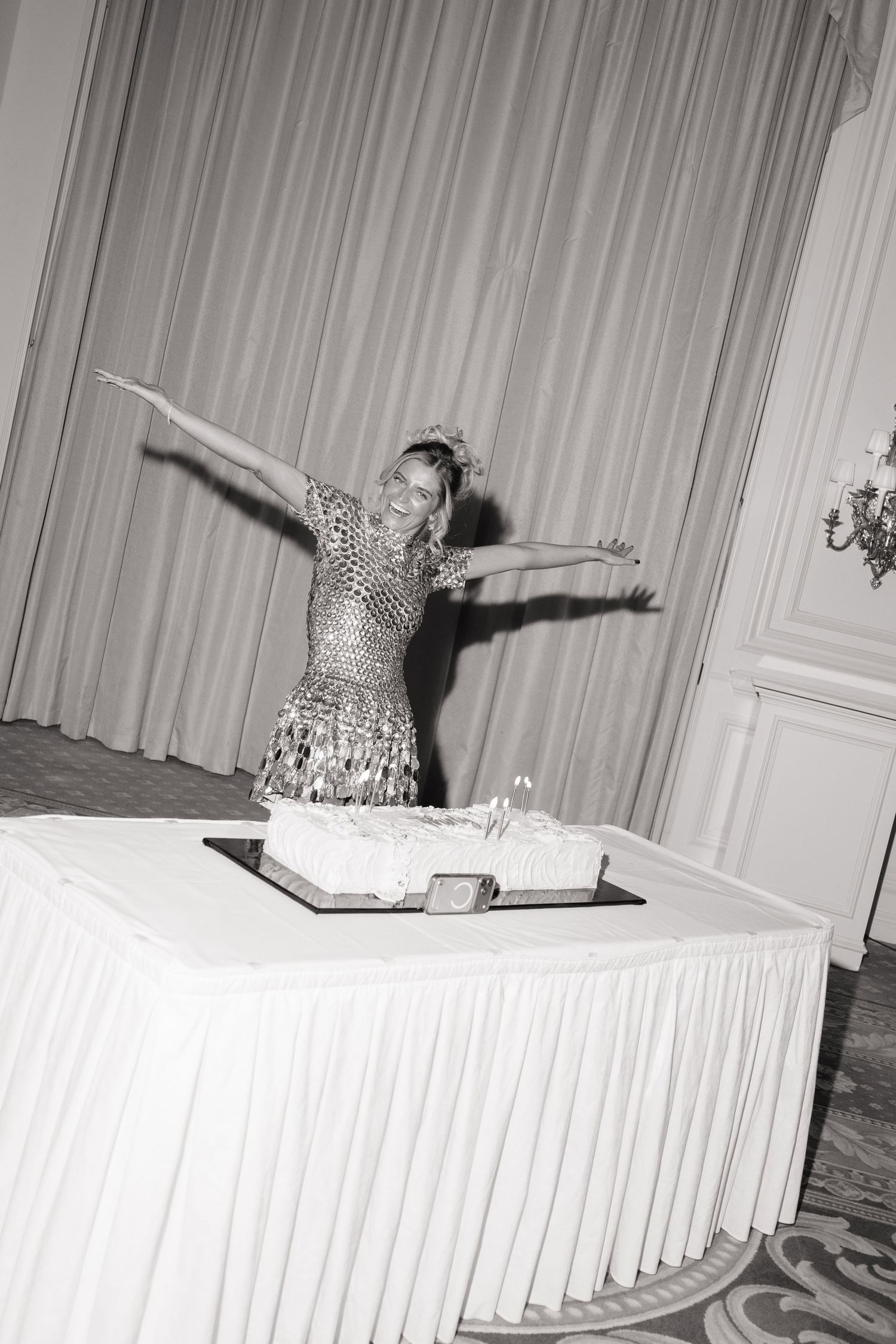 Woman in sequined dress poses with arms outstretched beside a large decorated cake at an elegant event.