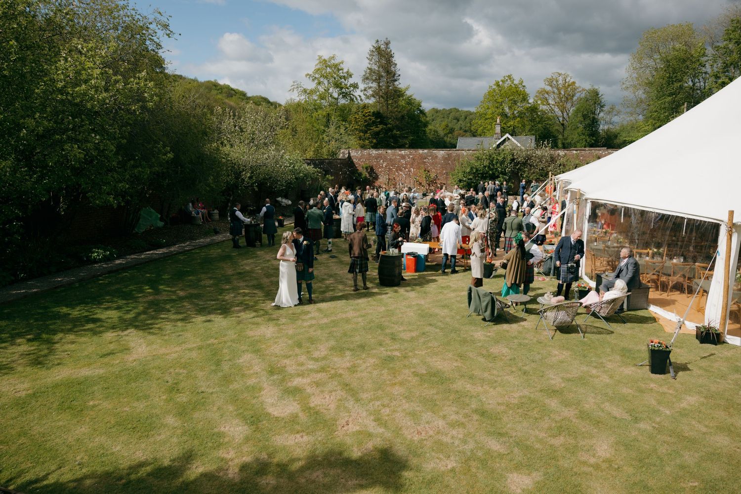 Aerial view of an outdoor wedding reception on a sunny day with guests gathered on a green lawn beside a white marquee tent.