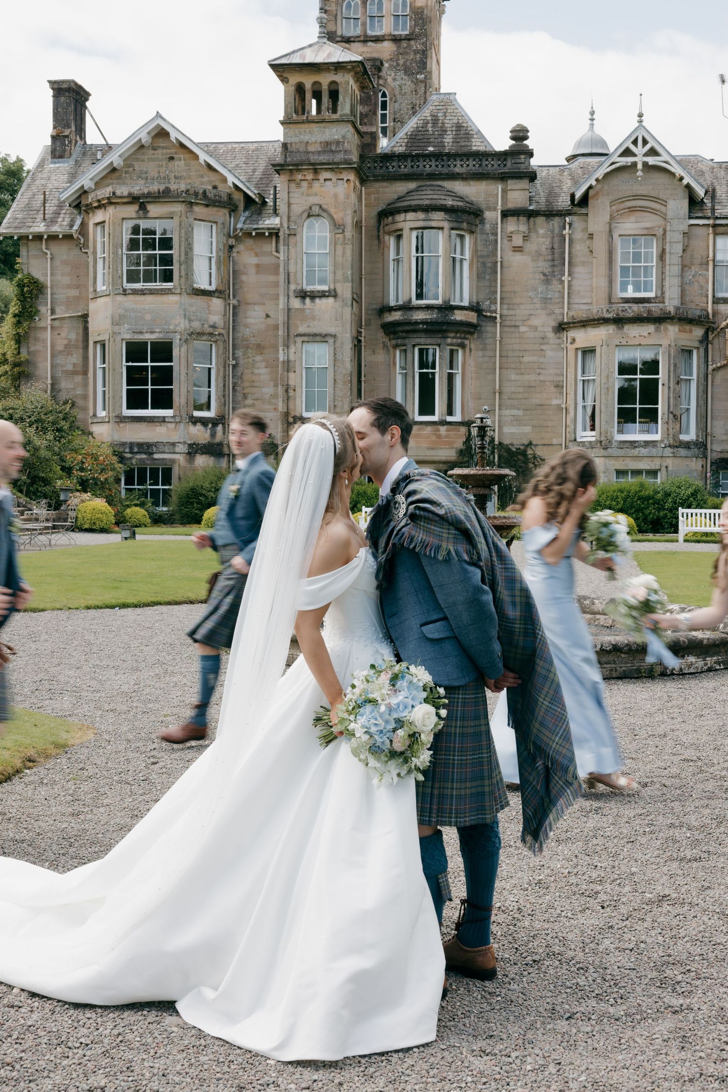 Bride and groom kiss in grounds of grand Scottish stone manor house with wedding guests nearby.