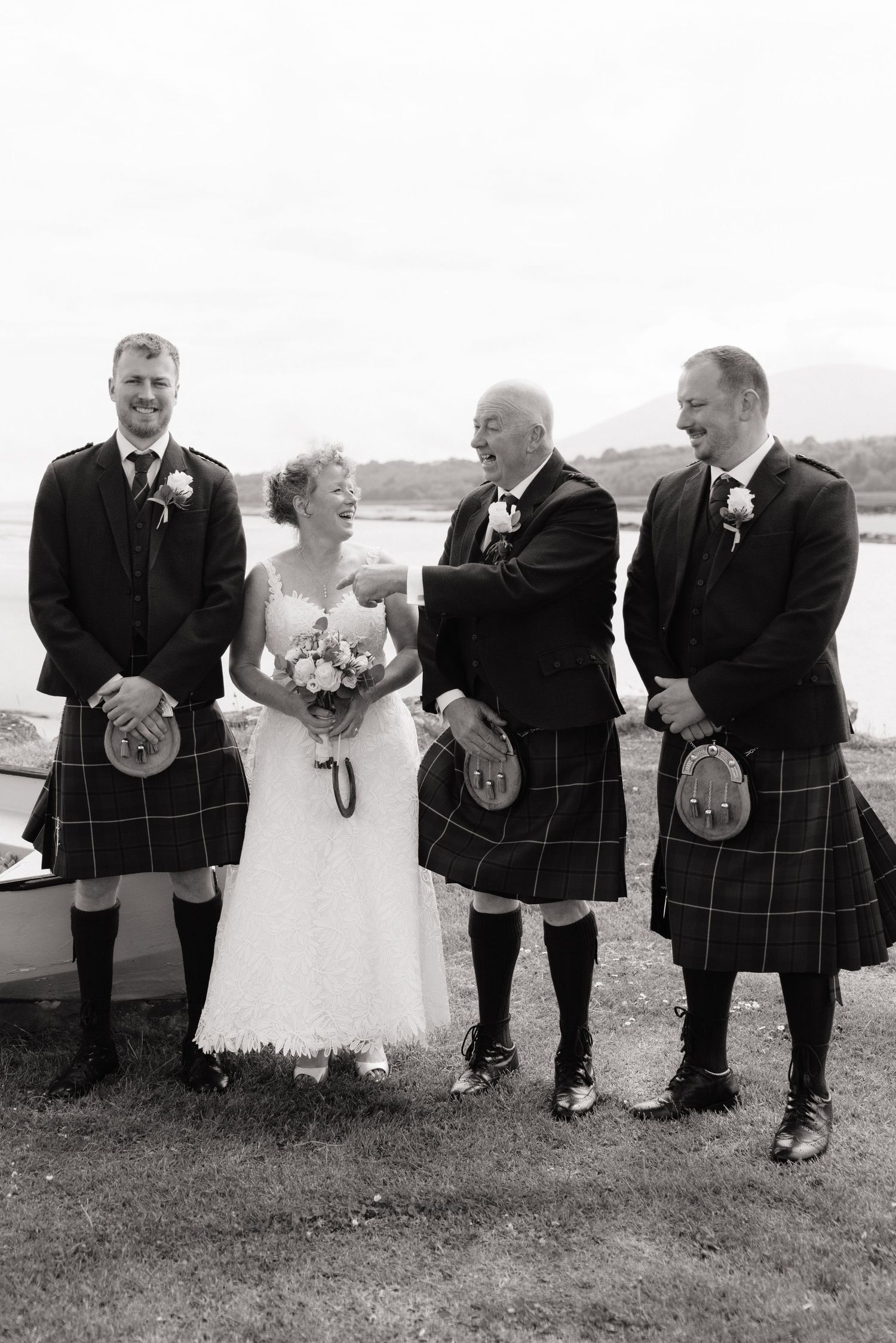 Black and white photo of a bride with three men in Scottish kilts laughing together outdoors near a loch.