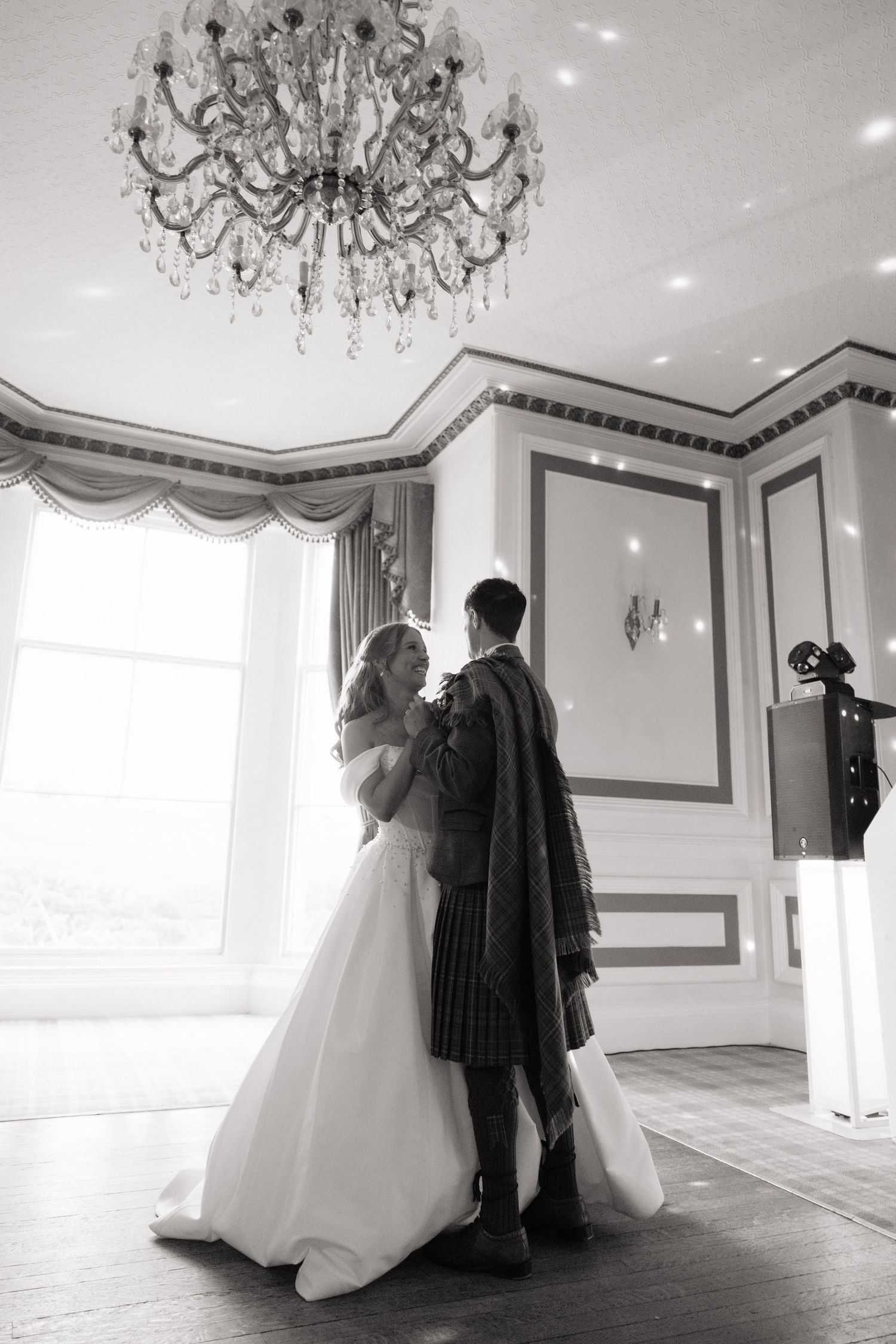 Black and white photo of bride and groom sharing first dance under crystal chandelier in elegant ballroom.